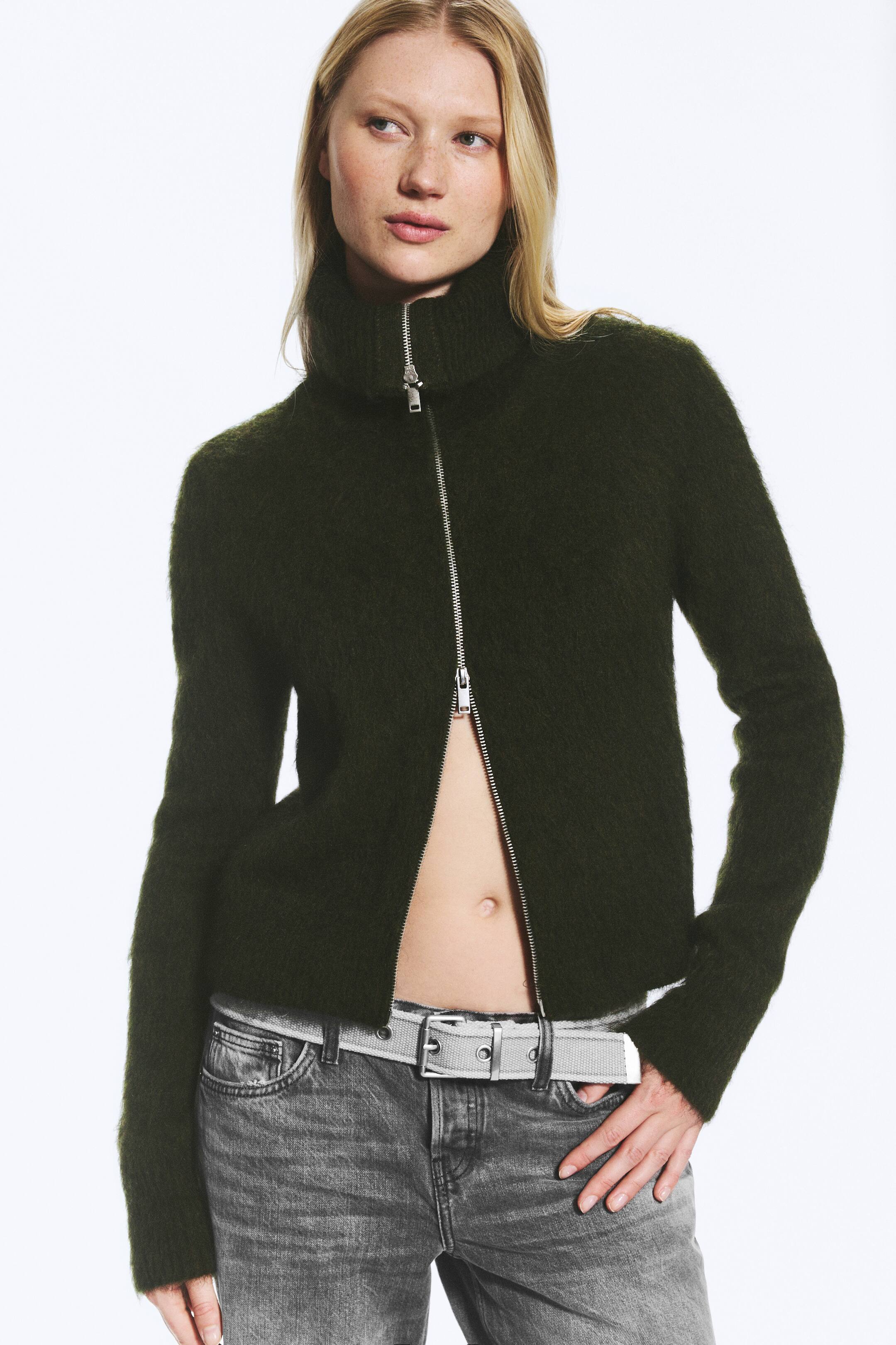 Grösseres Bild ansehen: Ein dunkelgrüner, flauschiger Cardigan aus Mohair-Mix wird vom Model getragen und verfügt über einen hohen Rollkragen sowie einen silbernen Zwei-Wege-Frontreissverschluss. Der Cardigan ist am Hals geschlossen und unten offen getragen, sodass der Bauchbereich sichtbar wird, kombiniert mit grau gewaschenen Jeans und einem hellgrauen Gürtel.
