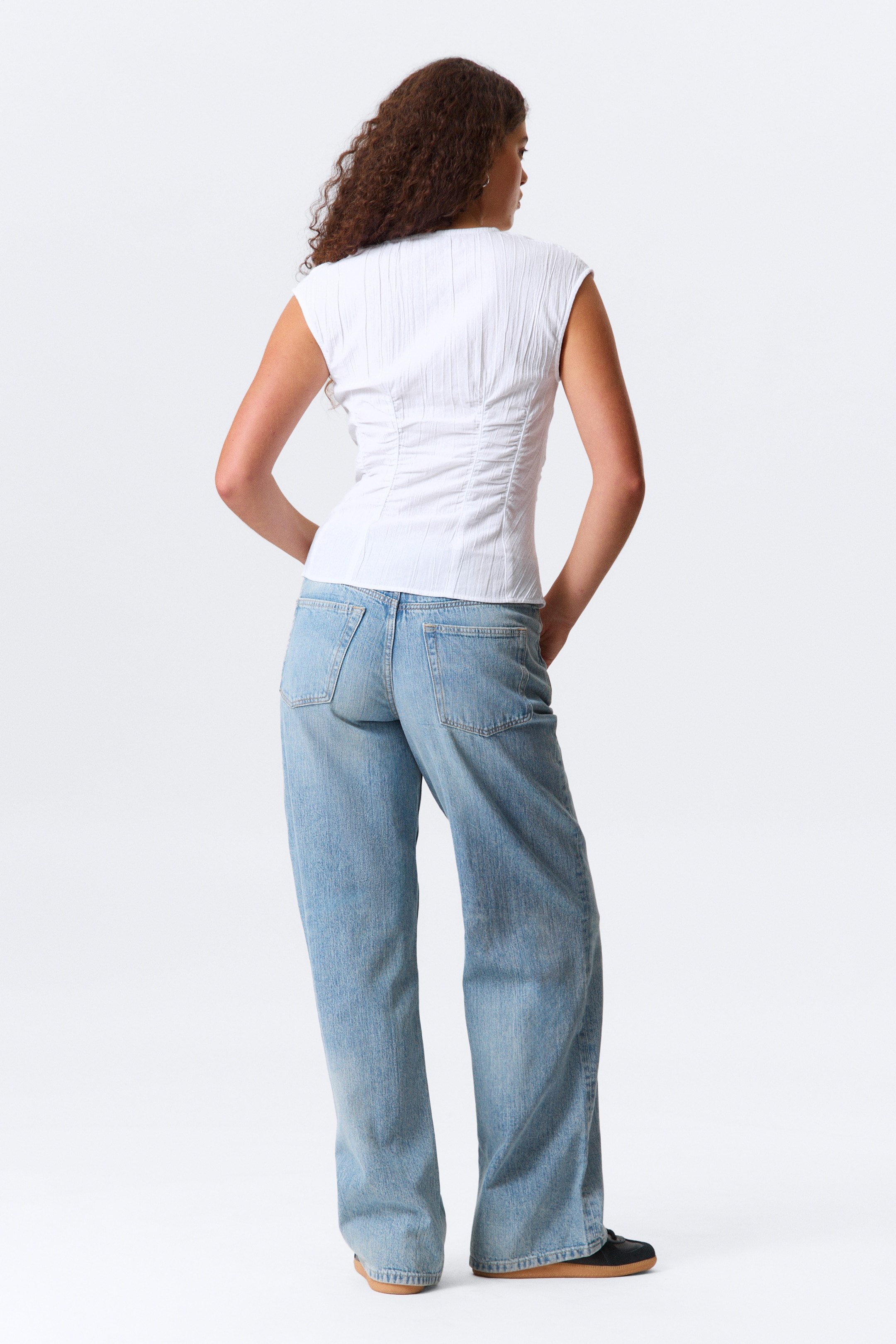 Visualizza immagine più grande: Un top bianco senza maniche, caratterizzato da pieghe testurizzate verticali e arricciature in vita per una silhouette aderente, è presentato da dietro. È abbinato a jeans a lavaggio chiaro e vestibilità rilassata.