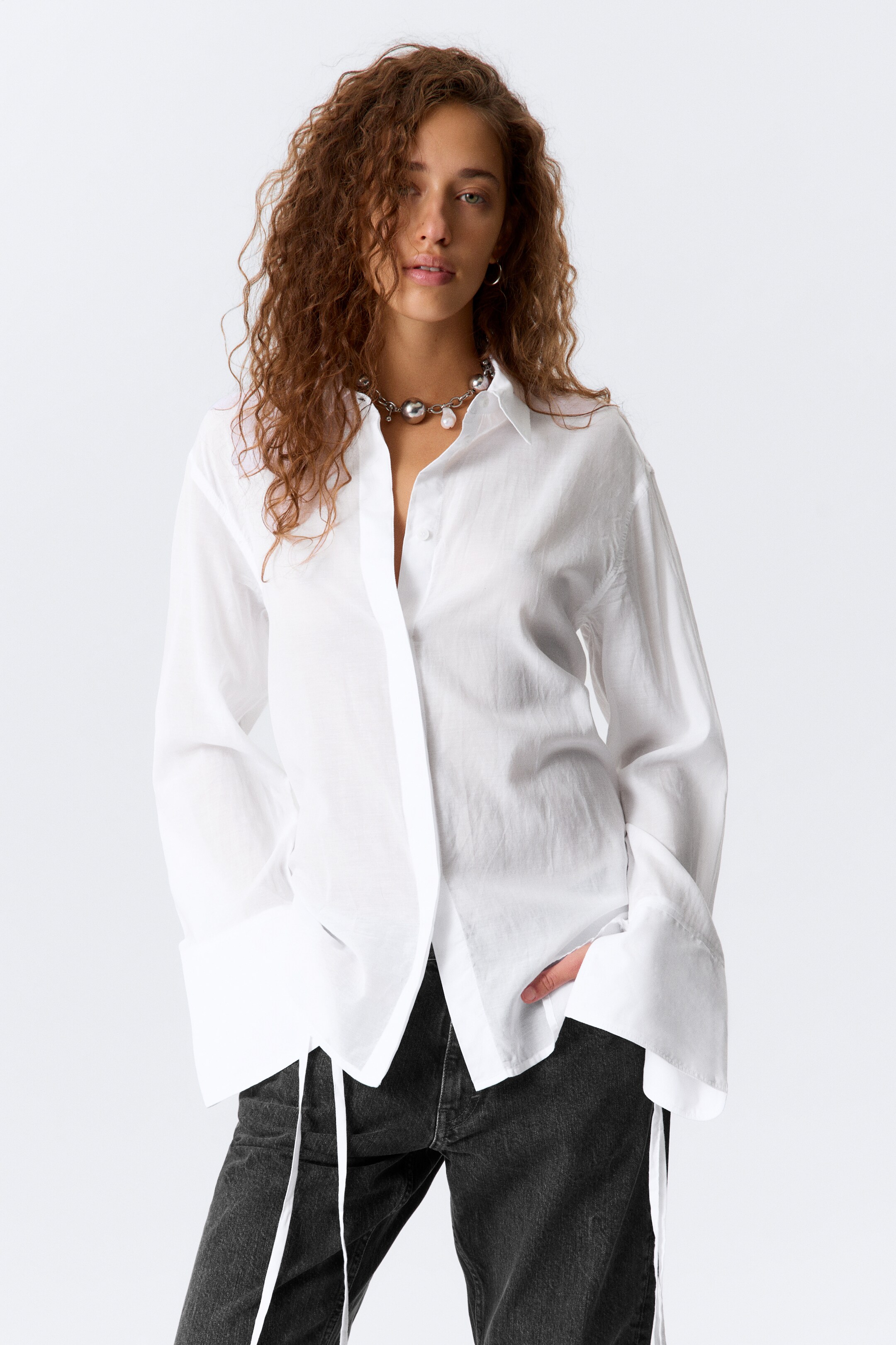 Ver imagem maior: Uma camisa branca oversized com botões está sendo usada, ligeiramente inclinada para a esquerda, apresentando uma gola pontiaguda e um placket frontal parcialmente desabotoado. Está a exibir mangas compridas com punhos largos e uma bainha assimétrica da qual pendem laços brancos.