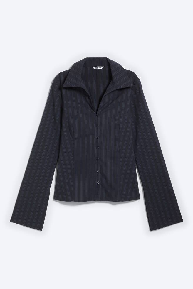 Een blouse met knoopsluiting wordt van voren gepresenteerd en toont een donker krijtstreeppatroon en een slank silhouet. Het beschikt over een diepe V-halslijn gecreëerd door een scherpe gespreide kraag, lange mouwen die wijd uitlopen bij de manchetten, en sluit aan de voorkant met knopen. - 1