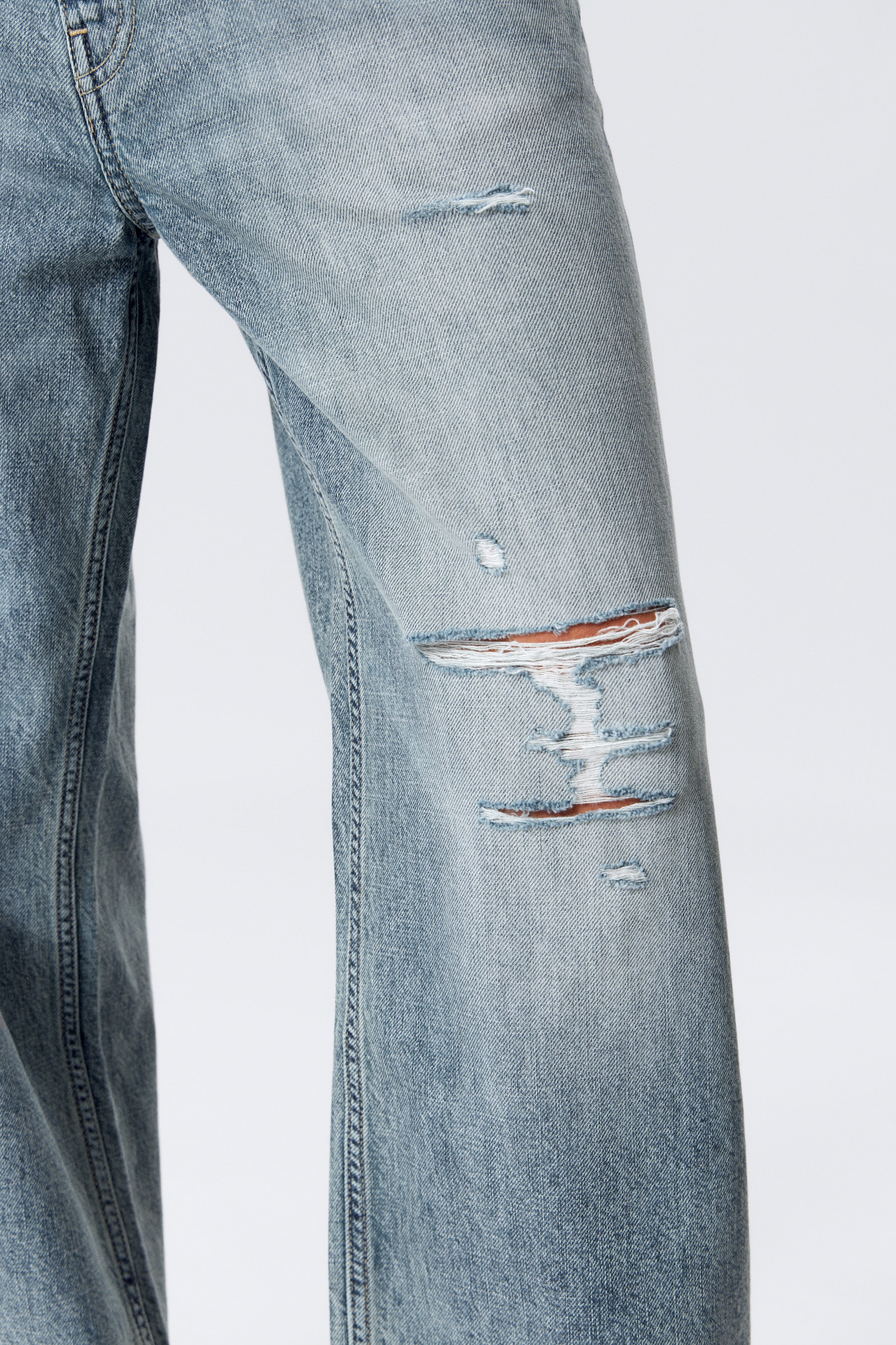 Grösseres Bild ansehen: Hellblaue Jeans aus Denim im Used-Look, mit mehreren Rissen und Löchern, die auffällig an der Vorderseite des Knies erscheinen und horizontale Fäden freilegen. Der Denim zeigt ein verblasstes Aussehen und markante vertikale Nähte verlaufen entlang der Beine.