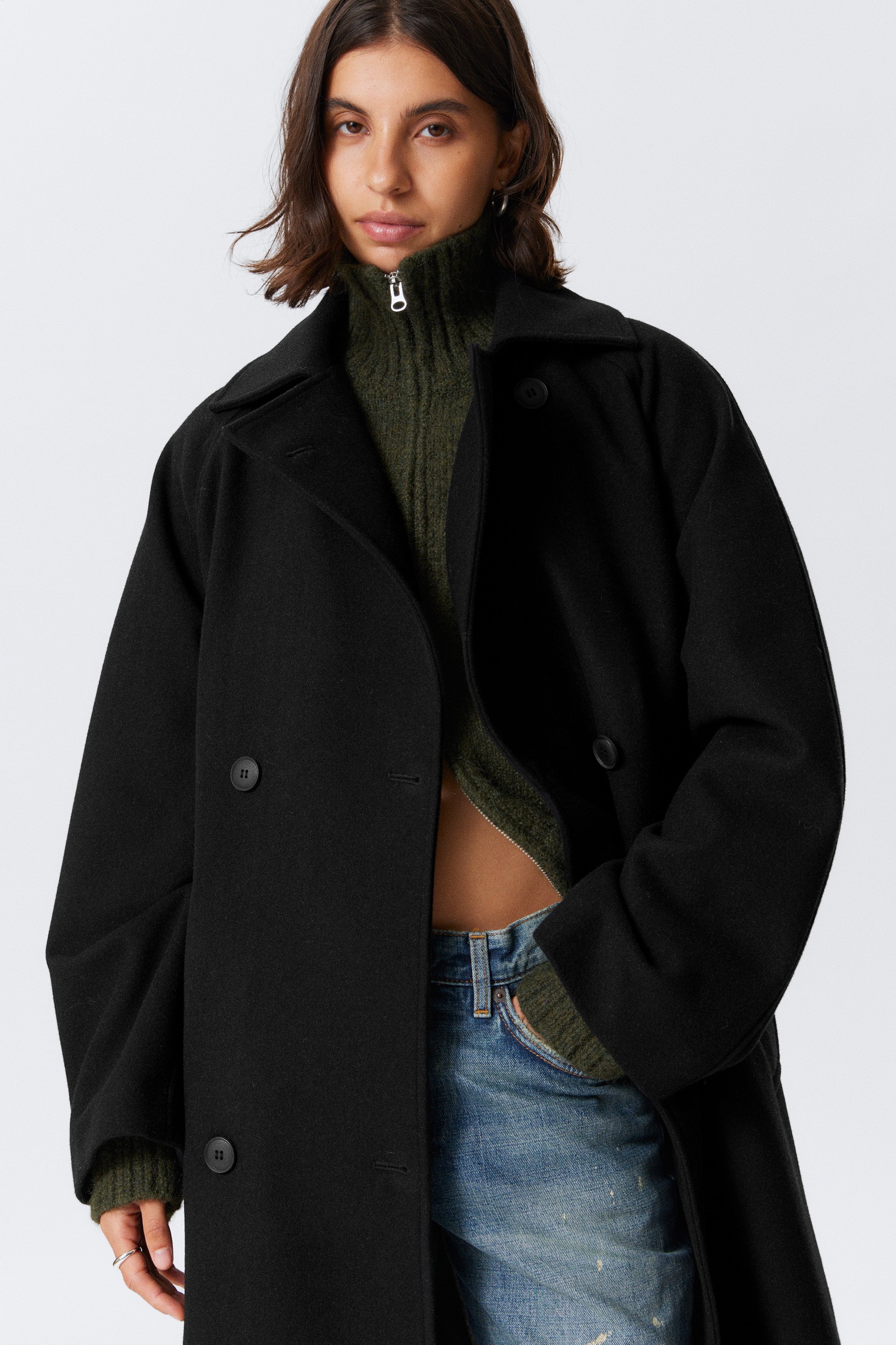 Visualizza immagine più grande: Un cappotto nero oversize doppiopetto viene indossato aperto dalla modella, con ampi revers a lancia e bottoni in tono. È abbinato a un maglione a collo alto a coste verde e jeans blu, con una delle mani della modella posizionata vicino alla parte inferiore frontale del cappotto.