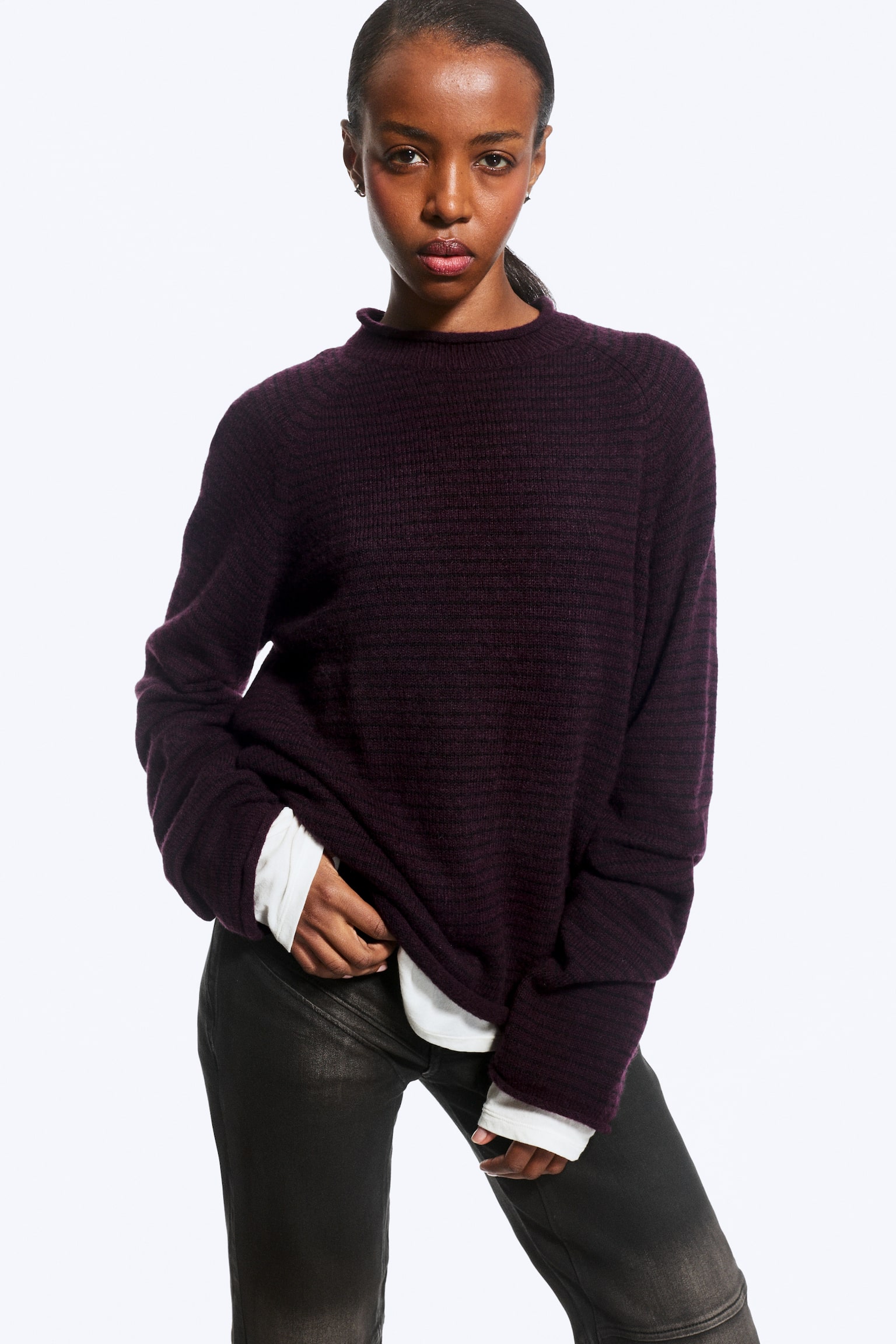 Un jersey de punto texturizado de color morado oscuro, con cuello chimenea y bordes sin rematar, lo lleva la modelo sobre una camiseta blanca de manga larga y pantalones oscuros. La modelo sujeta ligeramente de lado el bajo del jersey de corte holgado, resaltando su ajuste relajado y su estilo en capas. - 1