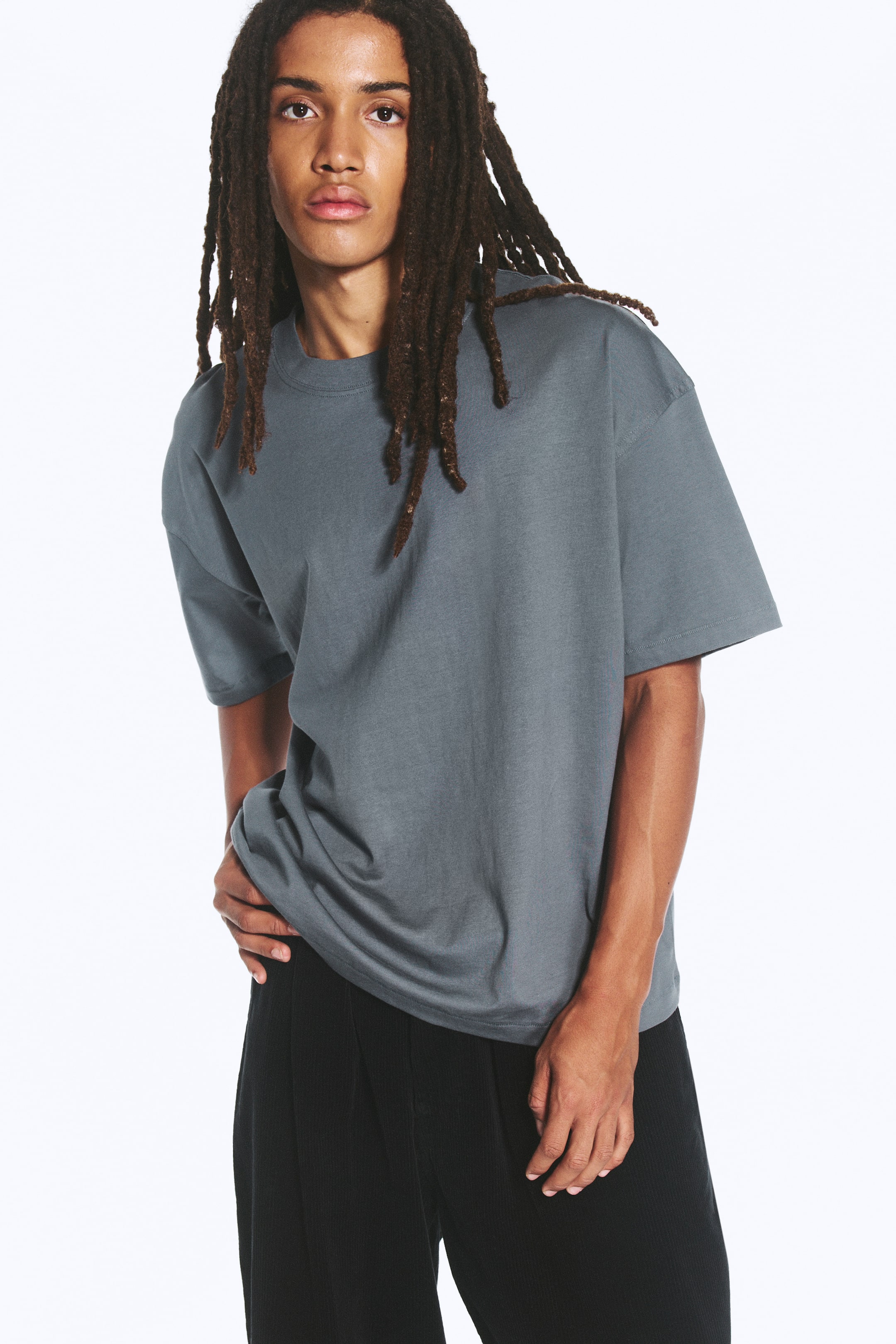 Προβολή μεγαλύτερης εικόνας: Ένα oversized boxy T-shirt σε σκούρο γκρι φοριέται από το μοντέλο, με στρογγυλή λαιμόκοψη, πεσμένους ώμους και φαρδιά μανίκια. Το T-shirt συνδυάζεται με μαύρο παντελόνι, ενώ το μοντέλο ακουμπά το ένα χέρι κοντά στο ισχίο.