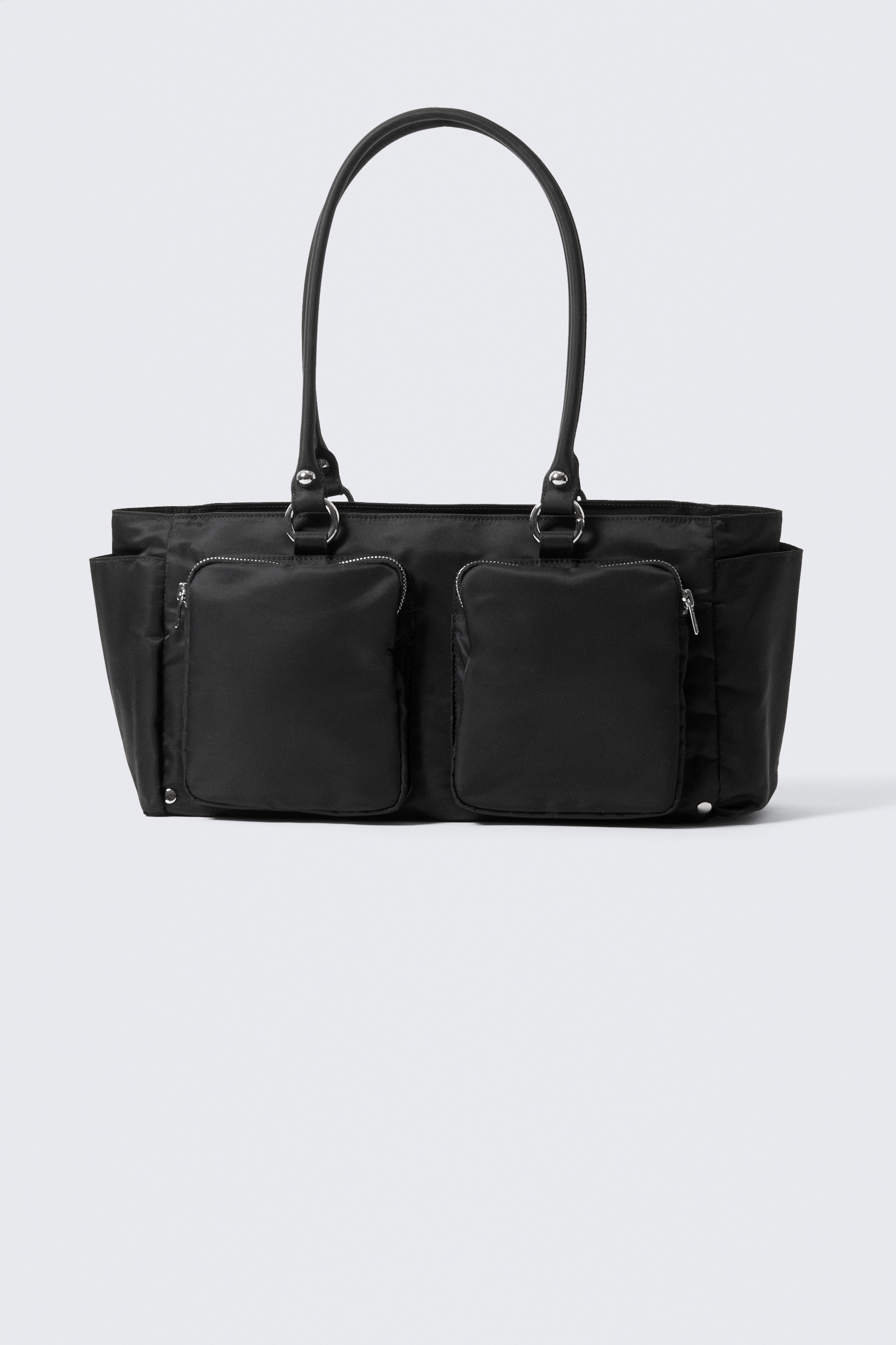 Agrandir l'image: Un sac fourre-tout noir est présenté de face, affichant deux poignées arquées fixées avec des ferrures de couleur argentée. Sa face avant est dotée de deux poches plaquées rectangulaires à fermeture éclair, et des poches ouvertes sont visibles sur ses côtés.