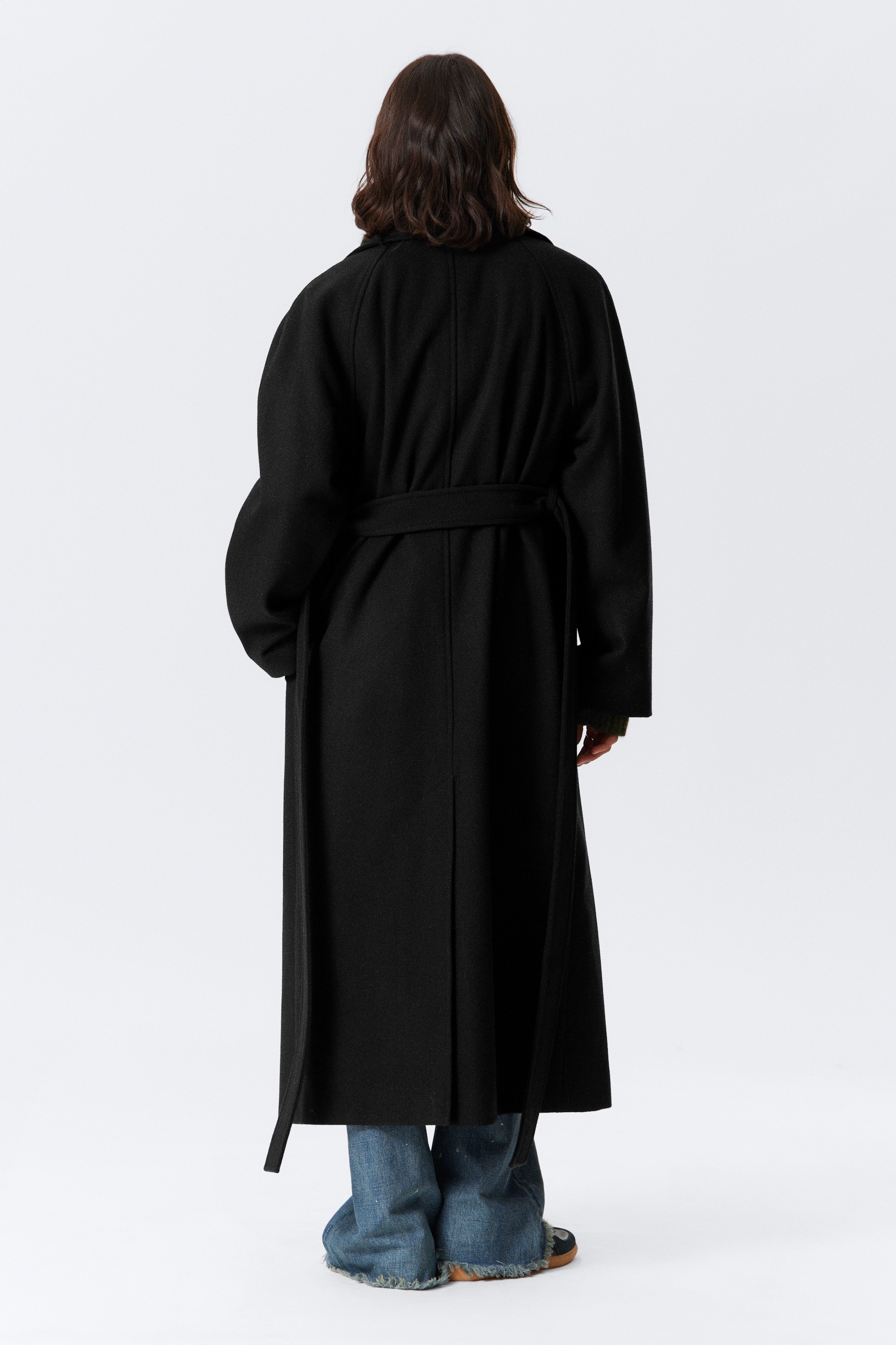 Visualizza immagine più grande: Un lungo cappotto nero oversize con maniche raglan è indossato dalla modella, vista da dietro. La cintura in tessuto auto è legata dietro, con le estremità che pendono liberamente.
