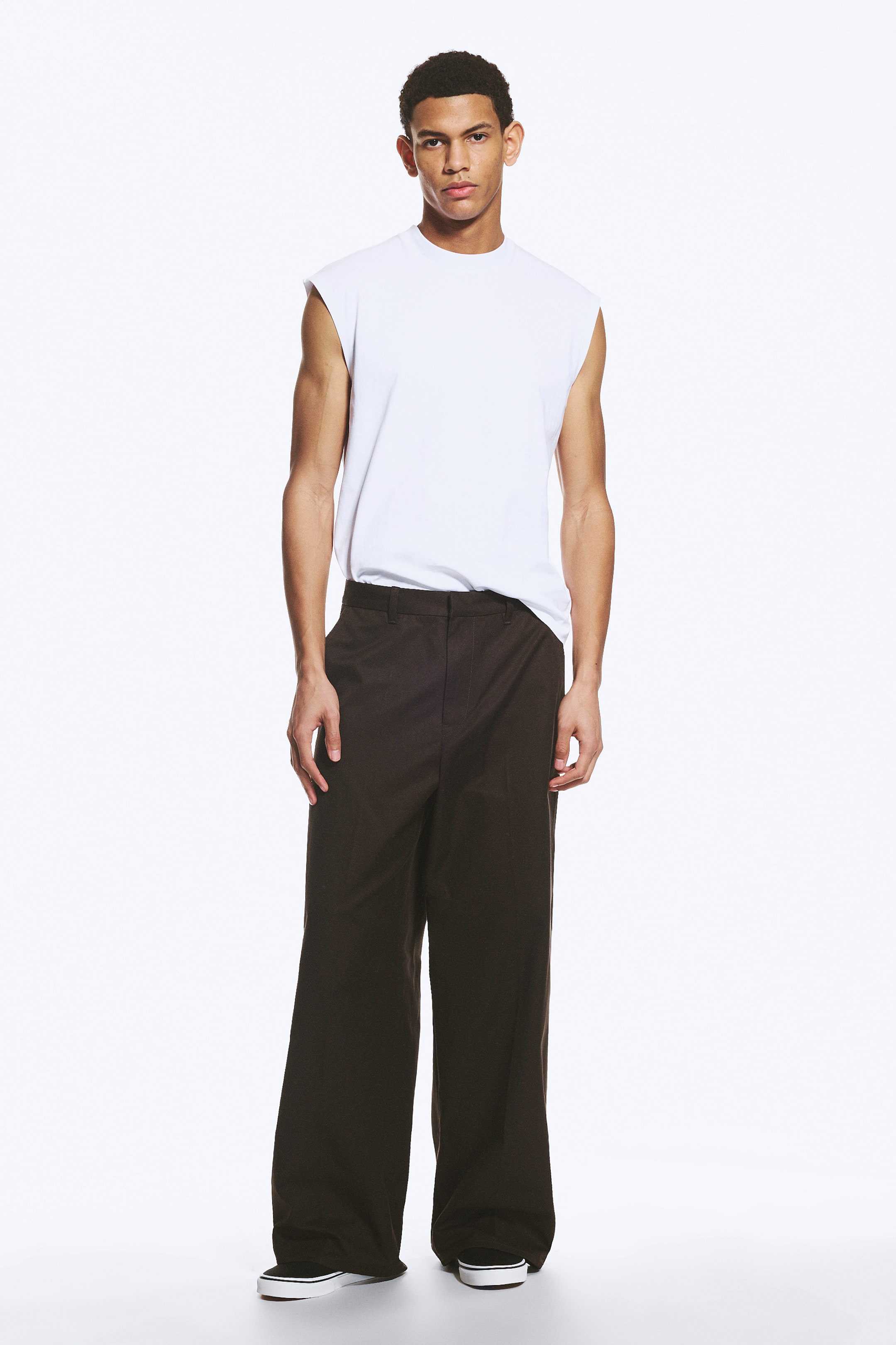 Ver imagem maior: Calças chino de cor castanho escuro, de corte largo, estão a ser usadas pelo modelo. As calças apresentam uma silhueta de perna larga, com vincos prensados a descer pela frente de cada perna e bolsos laterais inclinados visíveis nas ancas.