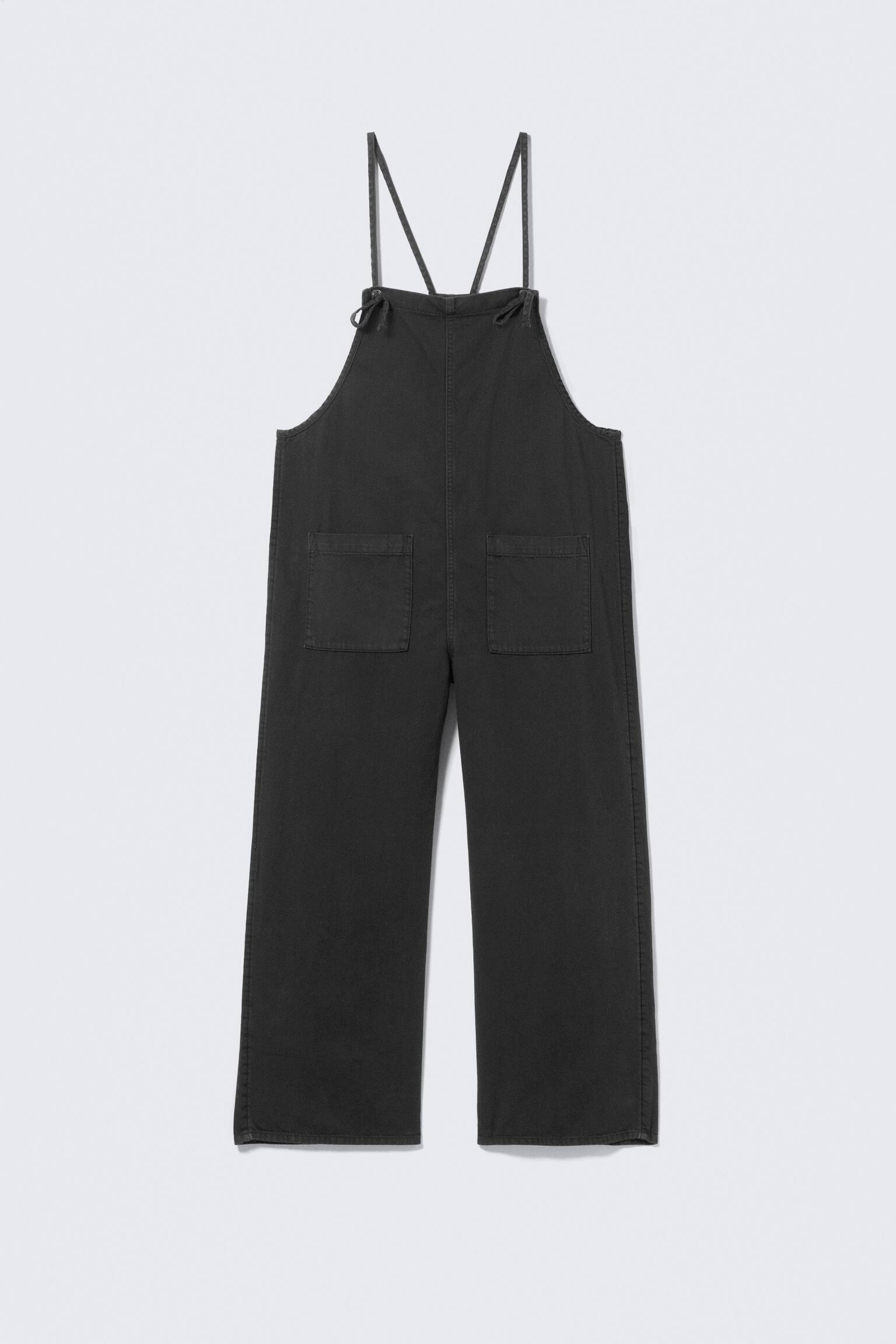 Dark grey dungarees are shown from a front view, featuring a bib front with thin tied shoulder straps. Two large rectangular patch pockets are visible on the bib, and the dungarees are wide-legged.