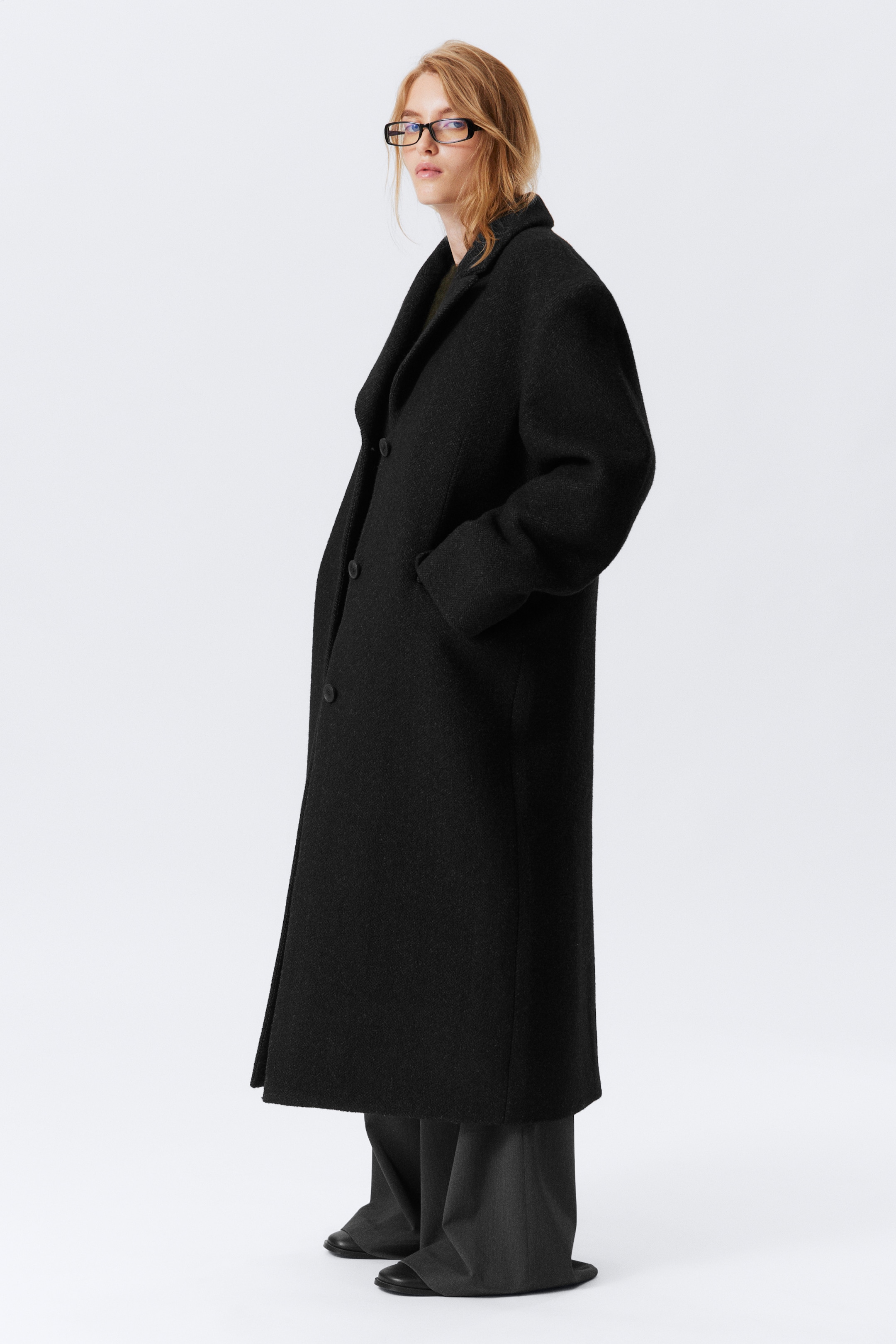 View larger image: A long, oversized black coat is being shown in a left profile view, featuring wide notch lapels, visible front buttons, and side pockets with one hand resting inside. The textured black coat is styled over dark wide-leg trousers, its hem reaching below the knees.