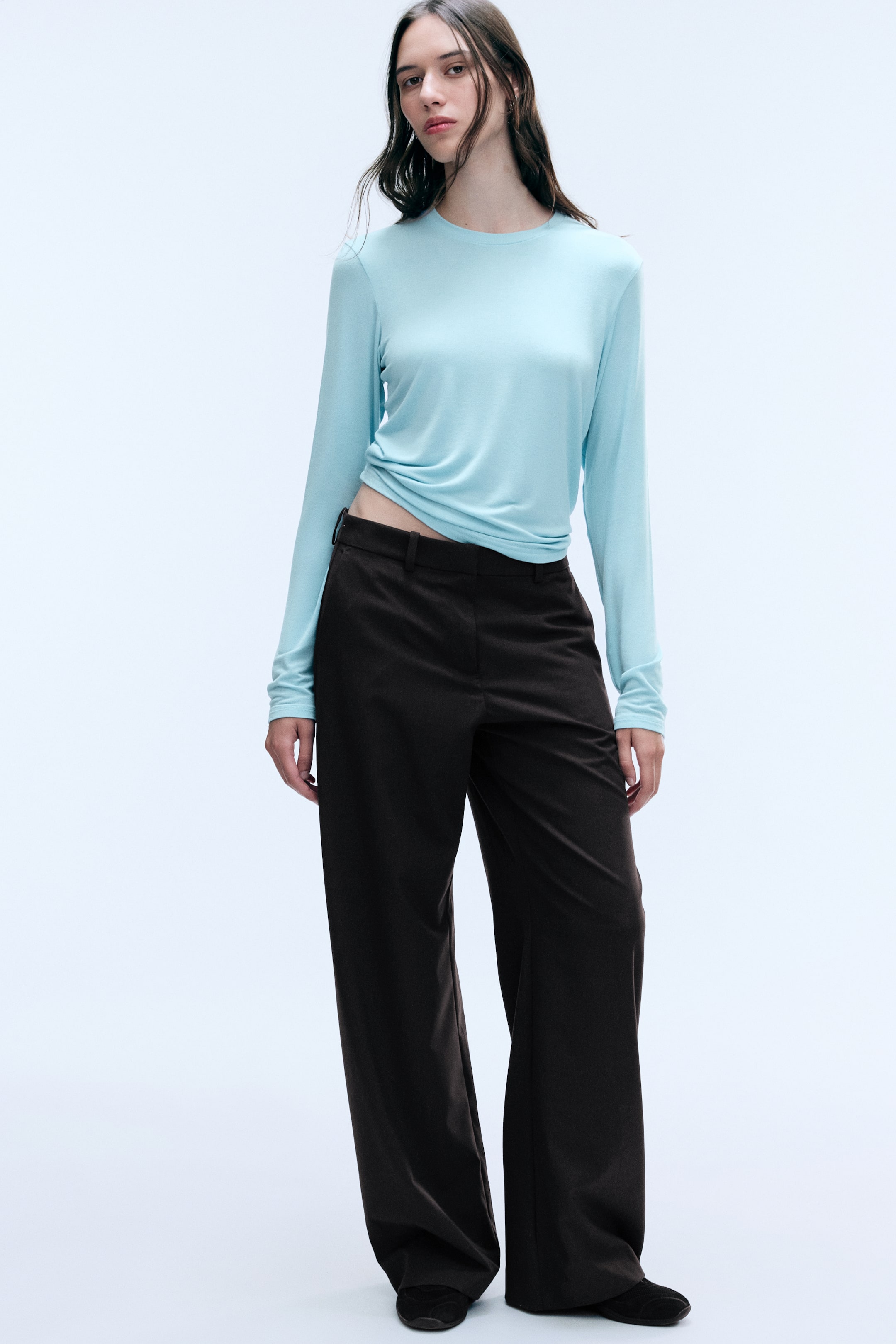 Größeres Bild anzeigen: Der Model trägt eine dunkelbraune Anzughose mit niedriger Taille, Gürtelschlaufen und lockerem, weitem Bein. Die Hose ist mit einem hellblauen Langarm-Top kombiniert, das an der Taille leicht gerafft ist.