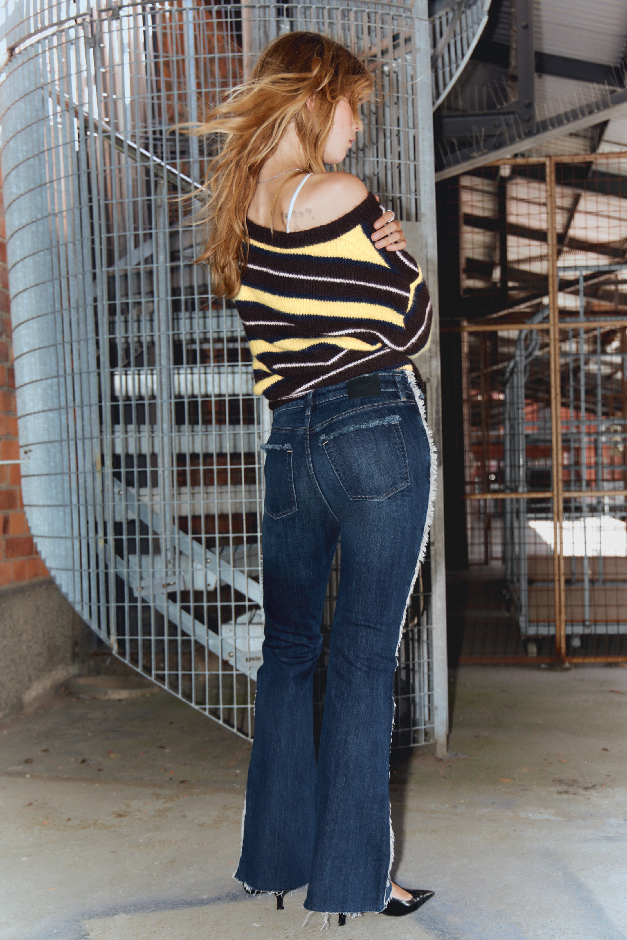 Größeres Bild anzeigen: Dunkel gewaschene Denim-Schlagjeans und ein gestreifter Strickpullover werden von hinten präsentiert. Die Jeans zeichnen sich durch ausgefranste Seitennähte und einen abgenutzten, rohen Saum aus, während der Pullover braune, gelbe und weiße horizontale Streifen mit einem schulterfreien Ausschnitt zeigt. Schwarze spitze Absätze werden mit den Jeans getragen.