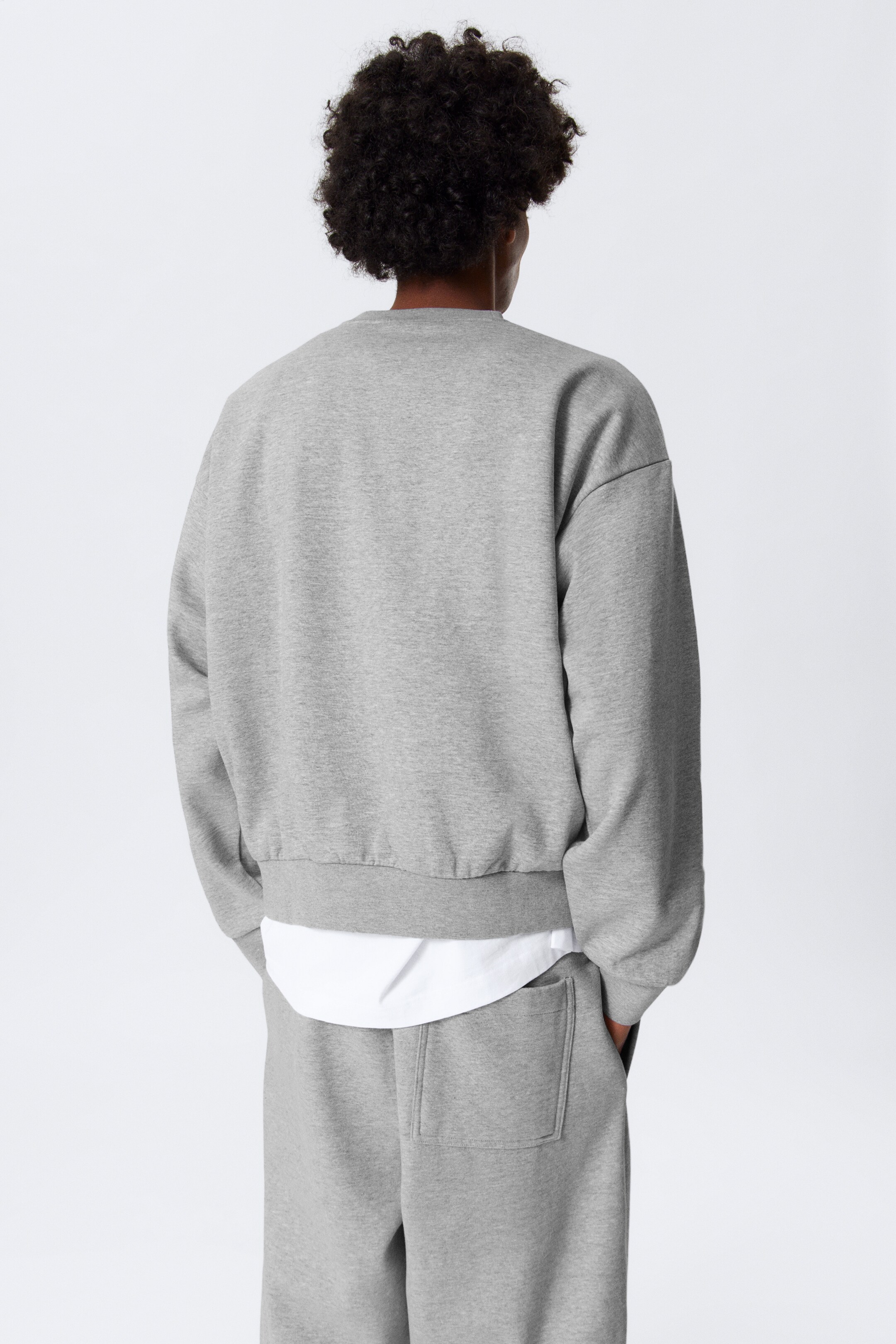 Größeres Bild ansehen: Ein graues Sweatshirt mit Rundhalsausschnitt wird von hinten gezeigt, es hat eine lockere Passform und einen gerafften Saum, darunter ist ein weißes Hemd sichtbar, das darunter getragen wird.
