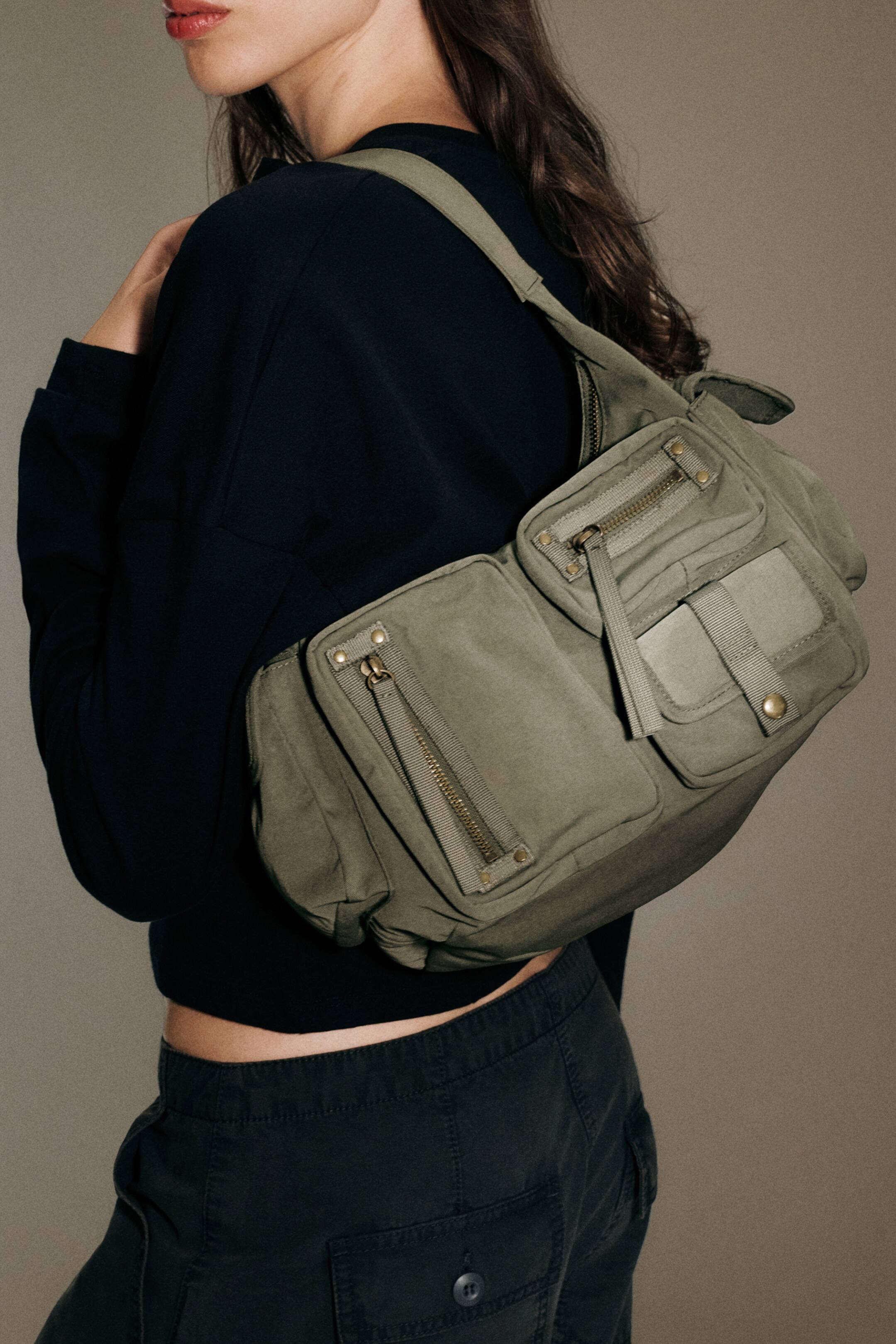 Ver imagem maior: Uma mala de ombro em lona verde-azeitona, com vários bolsos frontais com fechos de correr e botões de pressão, é usada ao ombro pela modelo. A mala compacta assenta de forma elegante sob o braço, combinada com uma camisola preta de manga comprida e calças escuras.