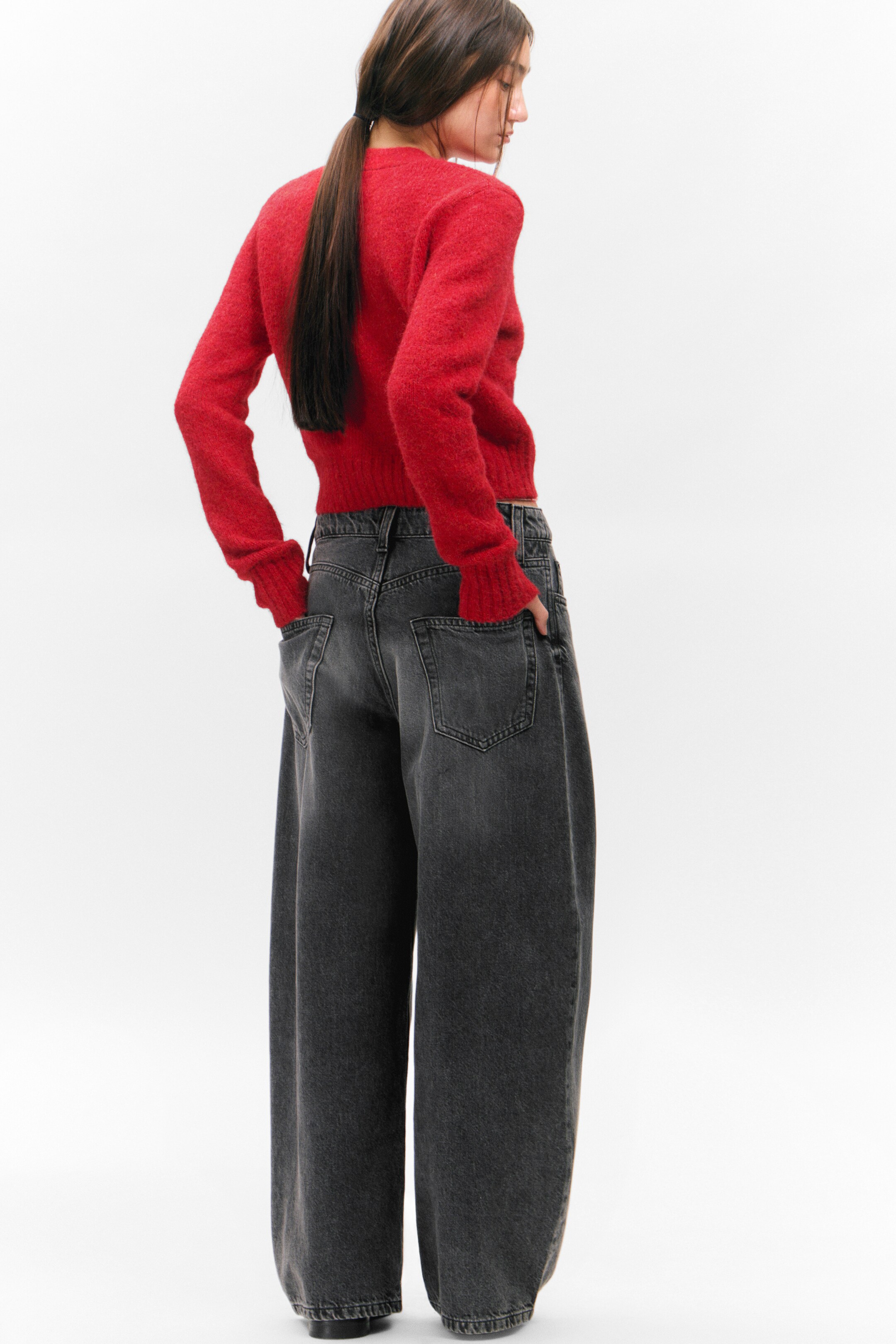 Agrandir l'image: Un cardigan rouge est porté par le mannequin, qui se tient debout les mains dans les poches d'un jean large et foncé. Le cardigan a des poignets côtelés et un ourlet côtelé, affichant un tricot texturé.