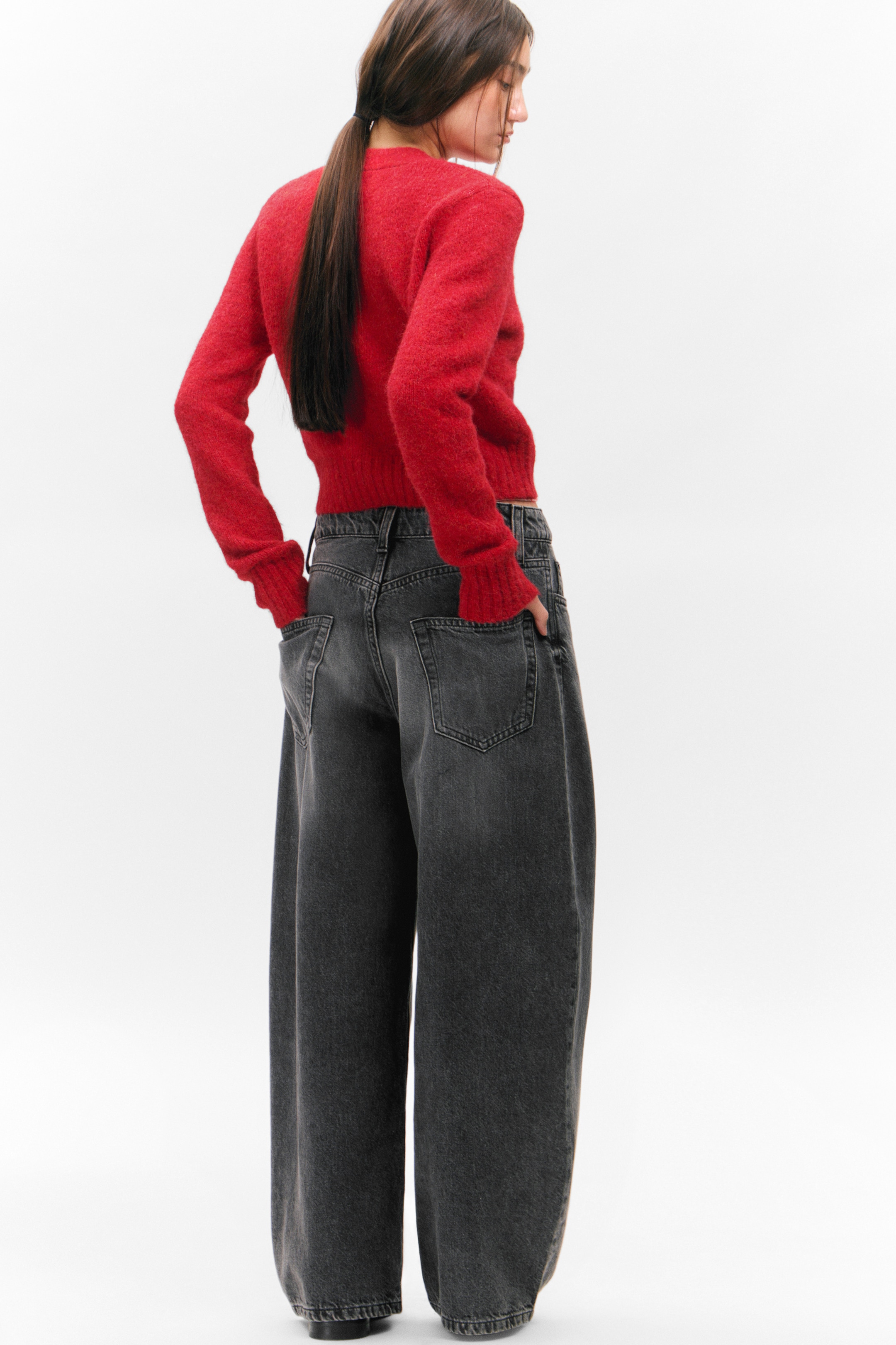 Größeres Bild ansehen: Ein roter Cardigan wird von dem Model getragen, das mit den Händen in den Taschen dunkler, weit geschnittener Jeans steht. Der Cardigan hat gerippte Bündchen und einen gerippten Saum, die ein strukturiertes Strickmuster zeigen.