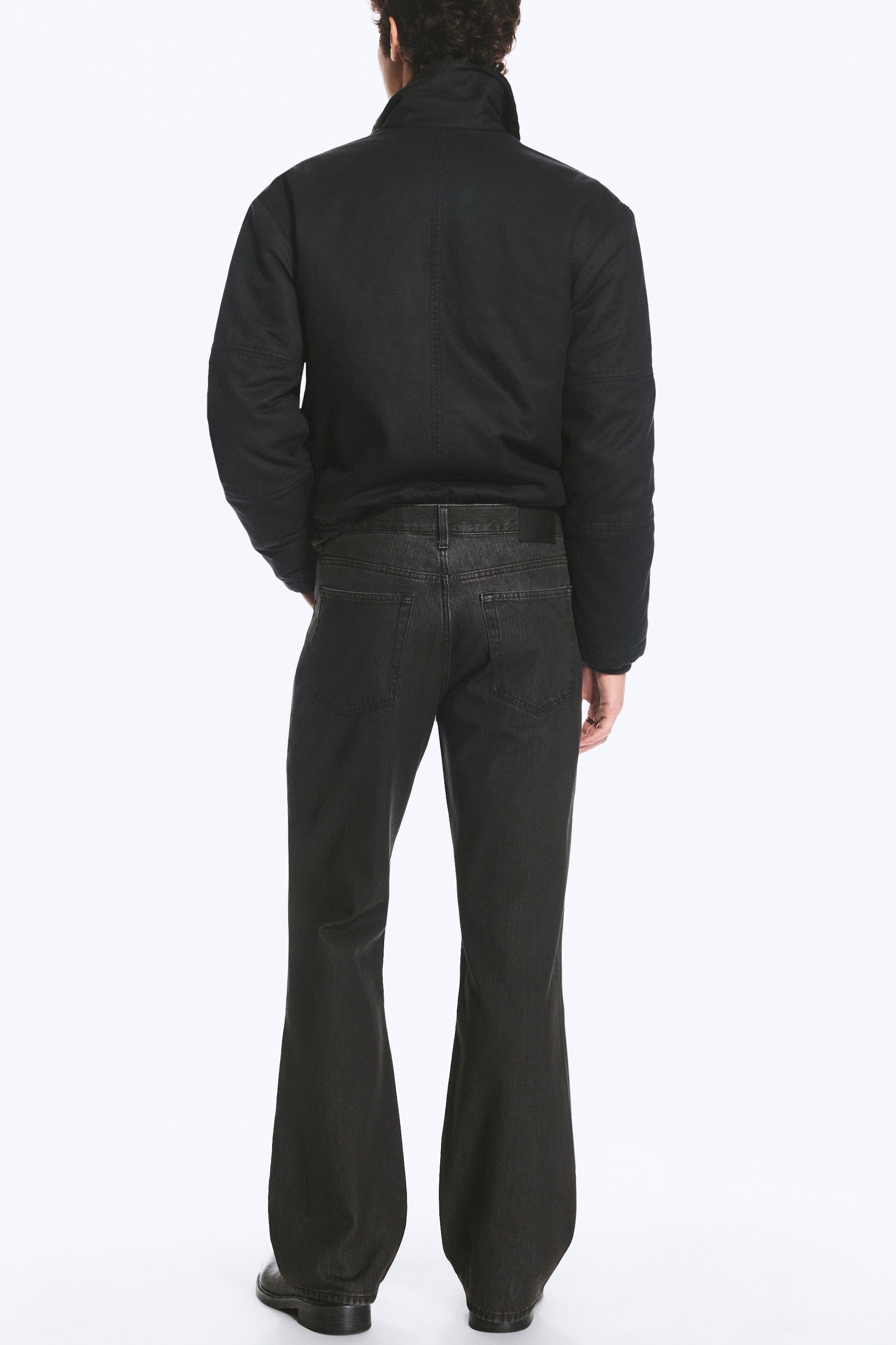Visualizza immagine più grande: Jeans in denim lavato nero, dal taglio ampio, con classico design a cinque tasche, indossati dalla modella e mostrati in una vista posteriore. I jeans sono abbinati a una giacca nera con colletto alto.