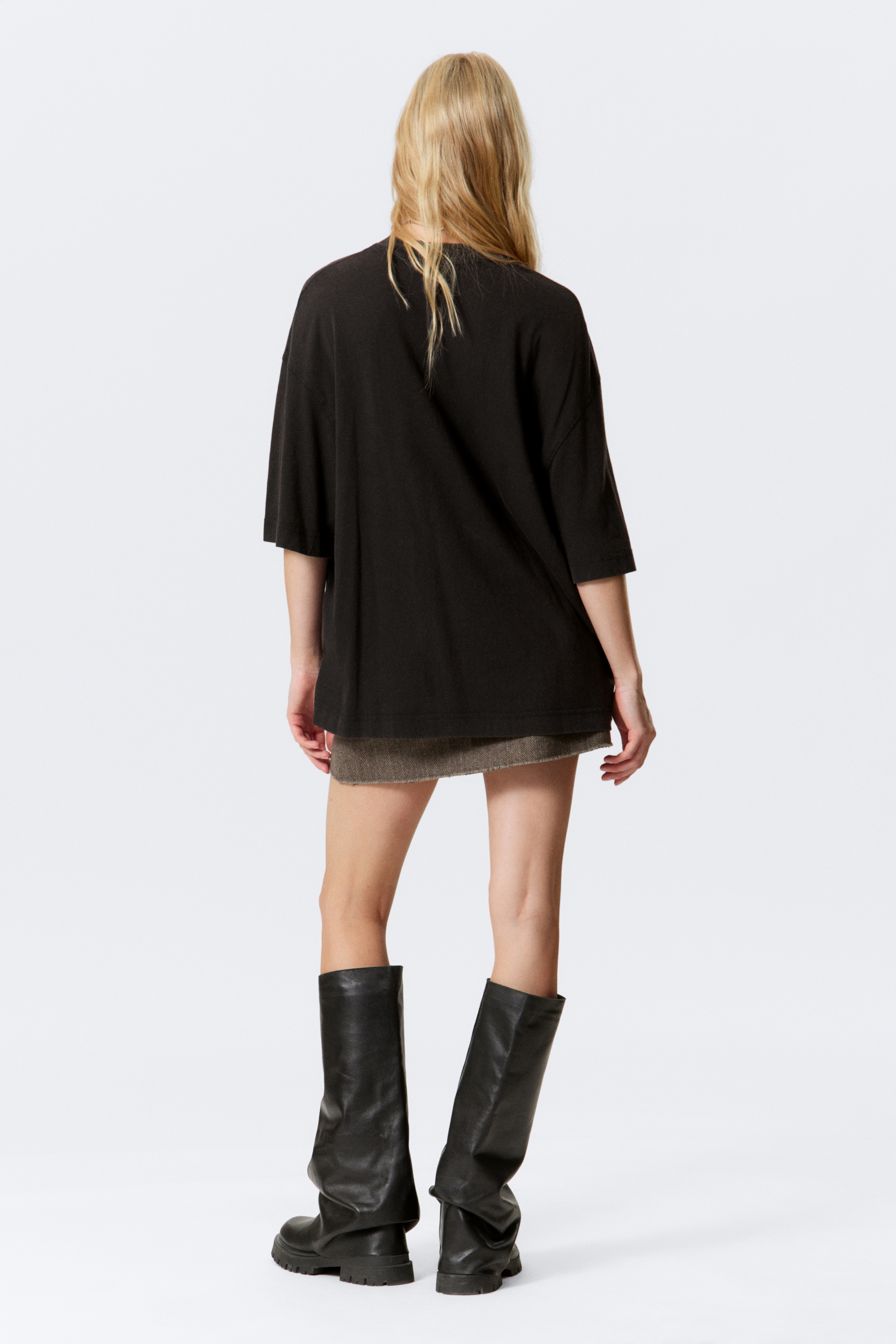 Agrandir l'image: Le T-shirt noir oversize est présenté de dos, avec une coupe décontractée et des manches courtes. Il est porté par-dessus une mini-jupe grise texturée, avec de hautes bottes noires à semelles épaisses également visibles.