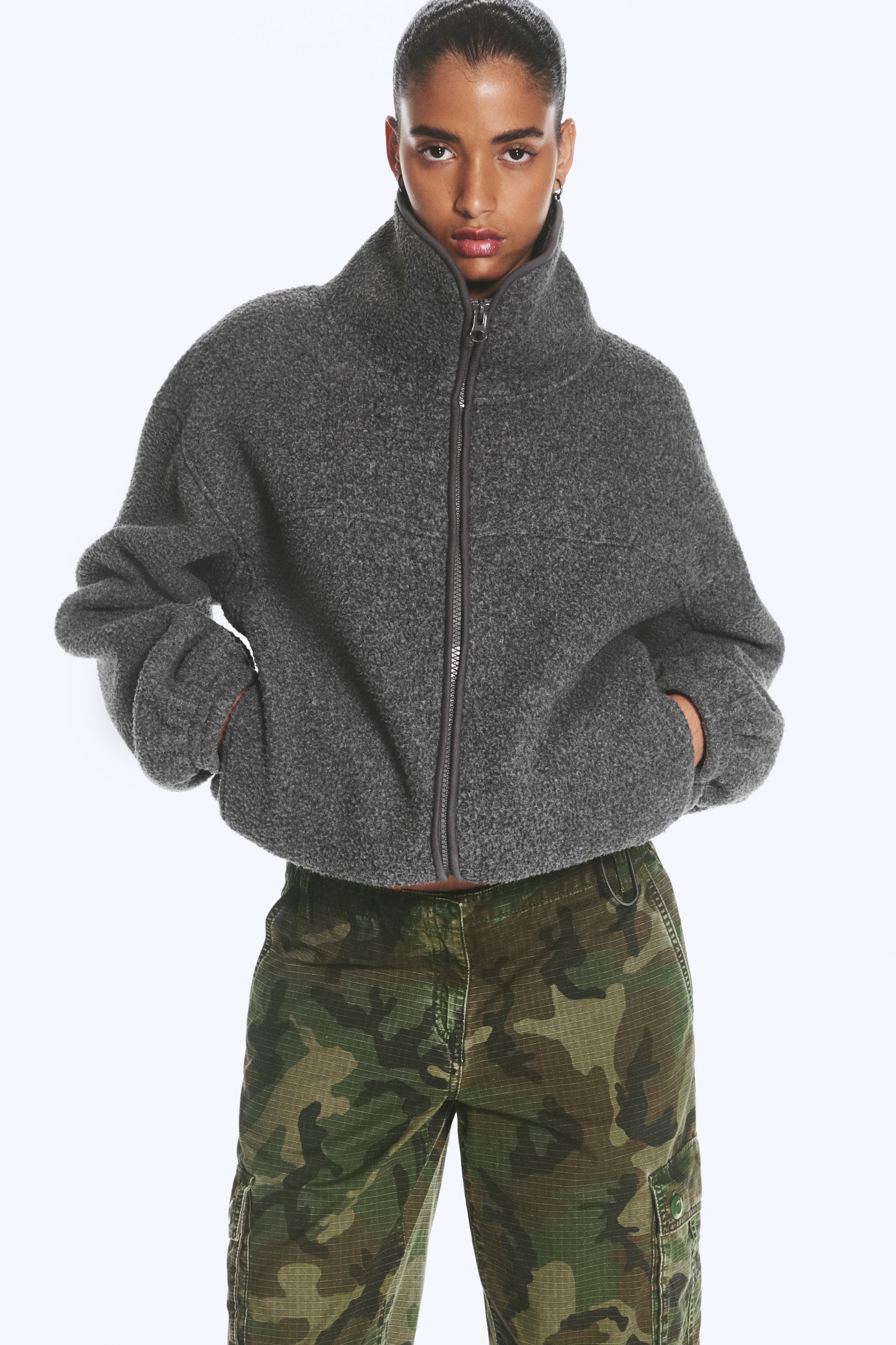 Größeres Bild anzeigen: Das Model trägt eine dunkelgraue Fleecejacke mit Stehkragen, die über eine strukturierte Fleece-Oberfläche, einen Frontreißverschluss und seitliche Taschen verfügt. Das Model hat die Hände in den Taschen und kombiniert die Jacke mit grünen Cargohosen im Camouflage-Look.