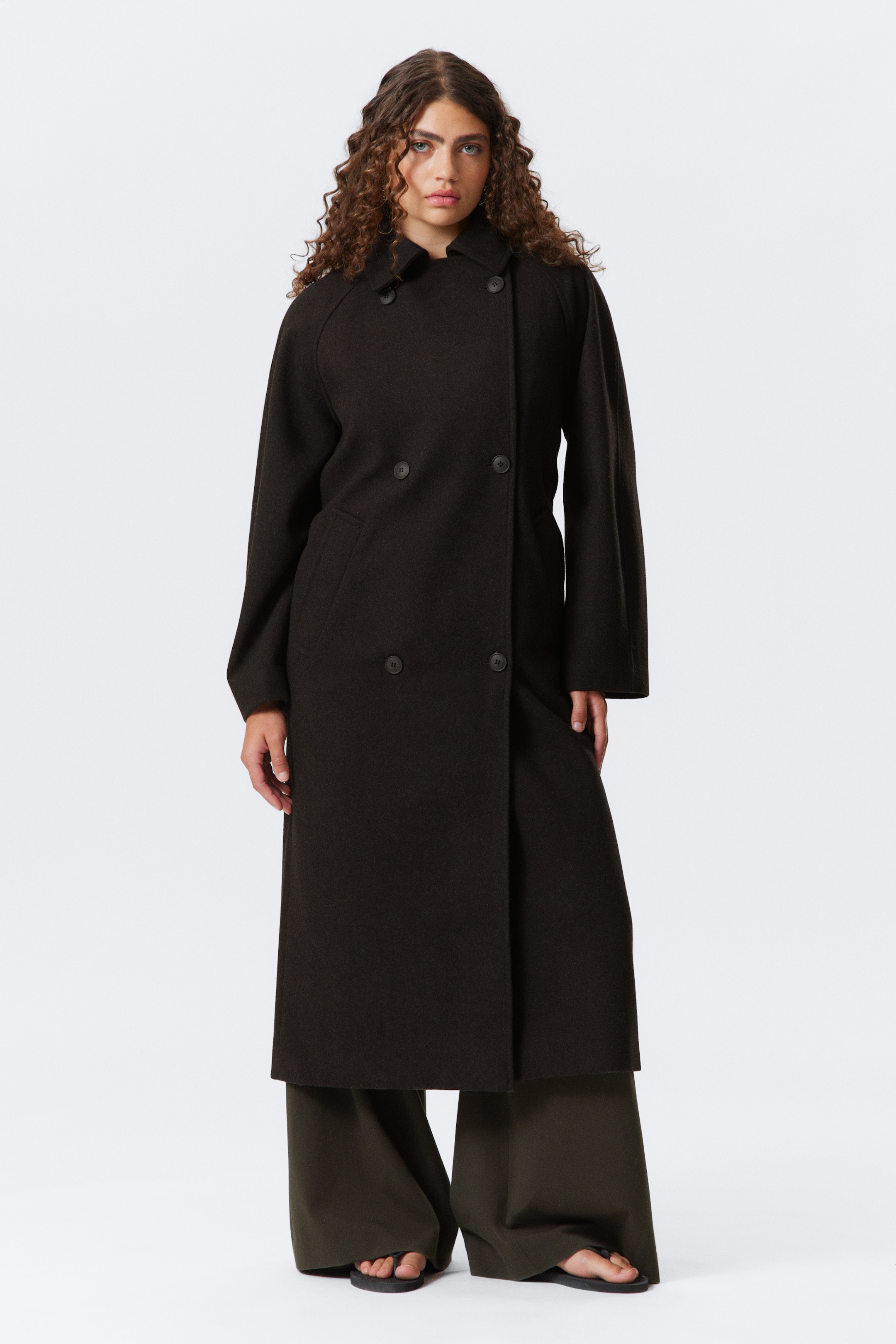 Agrandir l'image: Un manteau croisé marron foncé est présenté de face, avec une silhouette longue et décontractée qui s'étend sous les genoux. Il présente des manches raglan larges, un col haut boutonné, quatre boutons foncés et deux poches en biais.