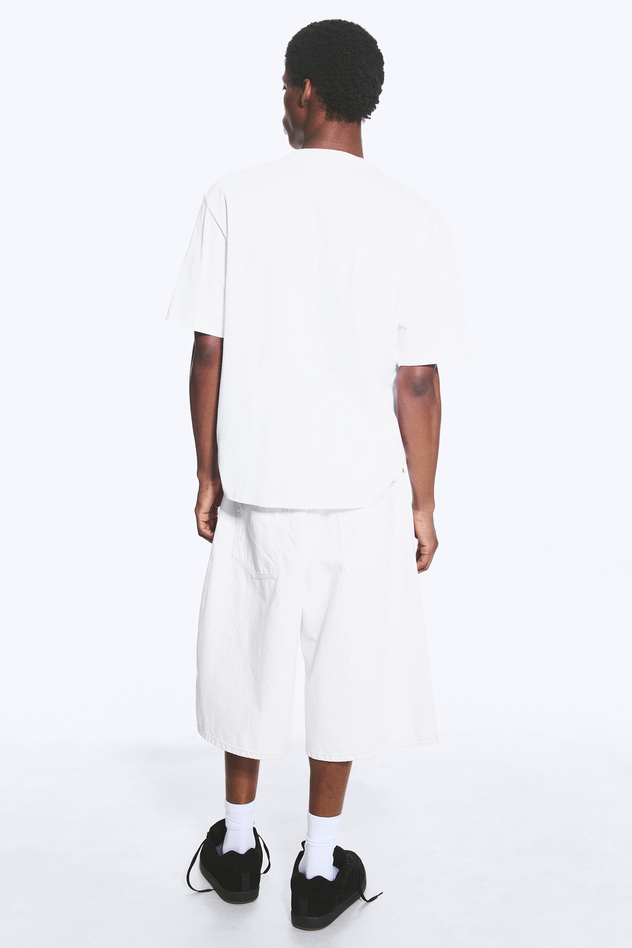 Ver imagen más grande: Una camiseta blanca de corte holgado con hombros caídos y dobladillo recto está siendo llevada por la modelo. La modelo está de espaldas al espectador, combinando la camiseta con unos pantalones cortos blancos.