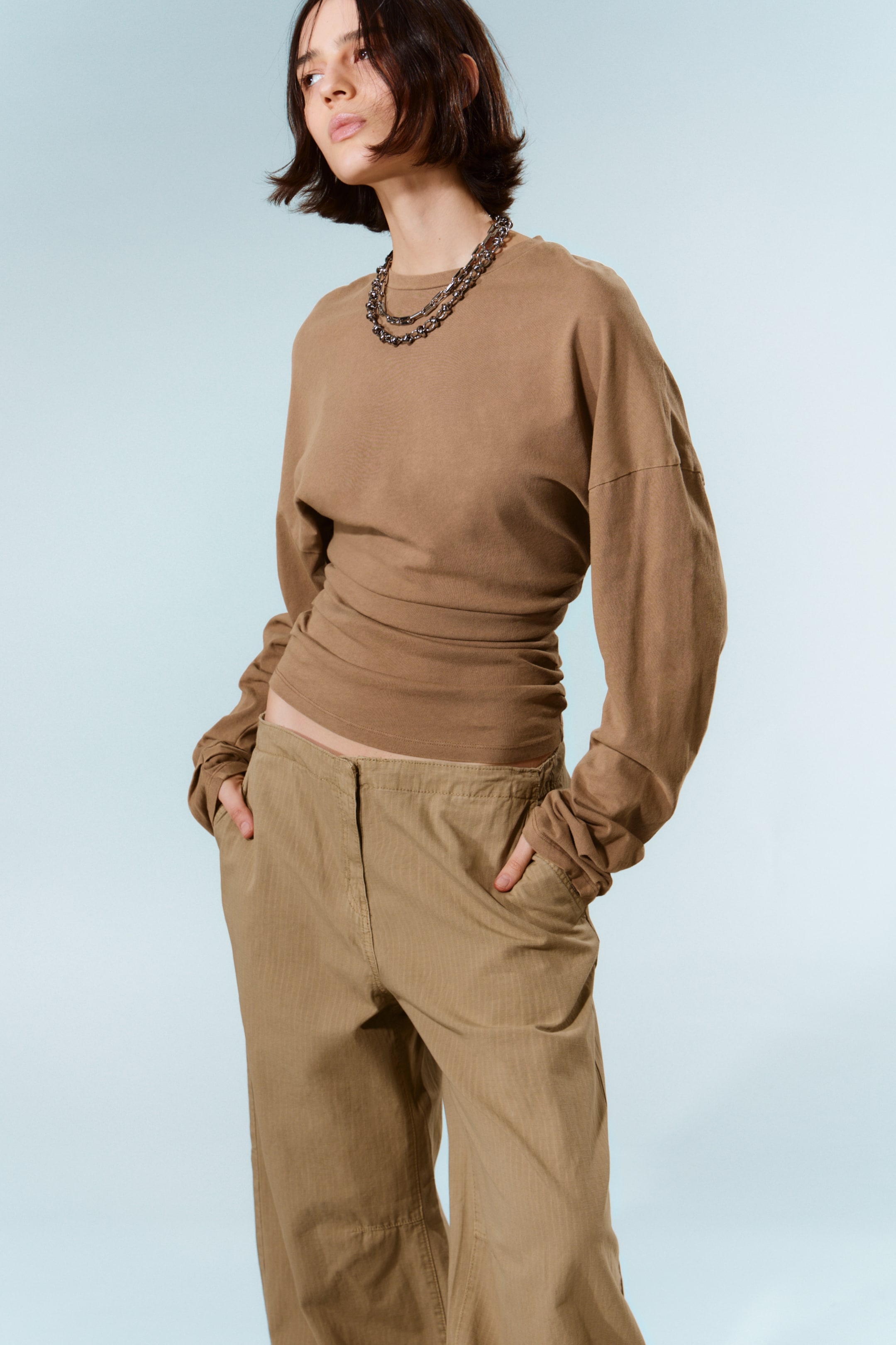 Zobacz większy obraz: Brązowy top z długim rękawem i okrągłym dekoltem charakteryzuje się luźnym krojem w okolicach ramion i rękawów oraz marszczonym, zwężanym efektem w talii. Na topie spoczywa warstwowy naszyjnik z metalowego łańcucha. Khaki spodnie z szerokimi nogawkami, ozdobione subtelnym pionowym wzorem w paski, bocznymi kieszeniami i płaskim przodem, doskonale uzupełniają top.