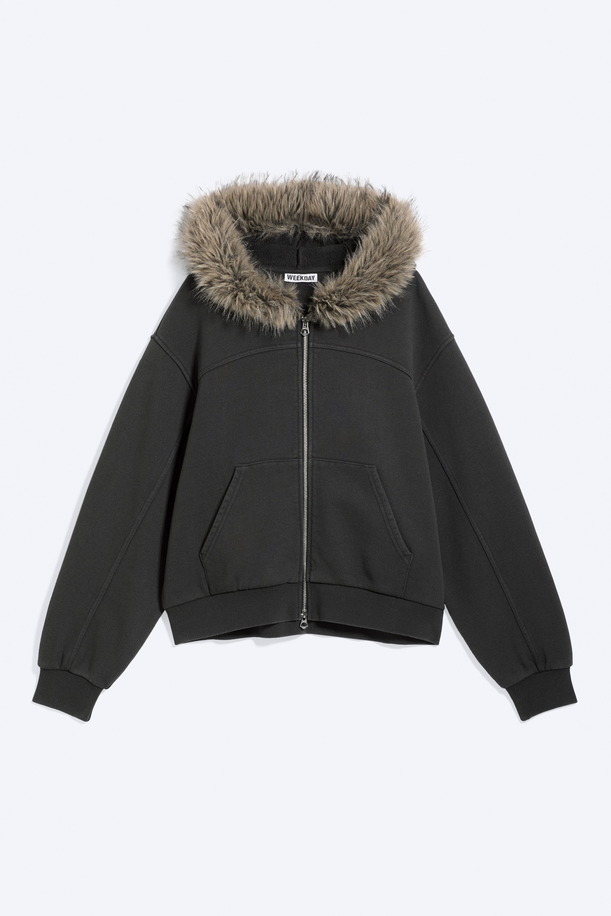 Un sweat à capuche zippé gris foncé, doté dâune capuche bordée de fausse fourrure claire, est porté par le mannequin, une main glissée dans une poche avant. Le sweat à capuche présente une fermeture à glissière frontale bidirectionnelle partiellement ouverte et des poignets côtelés, et il est associé à une jupe assortie gris foncé.