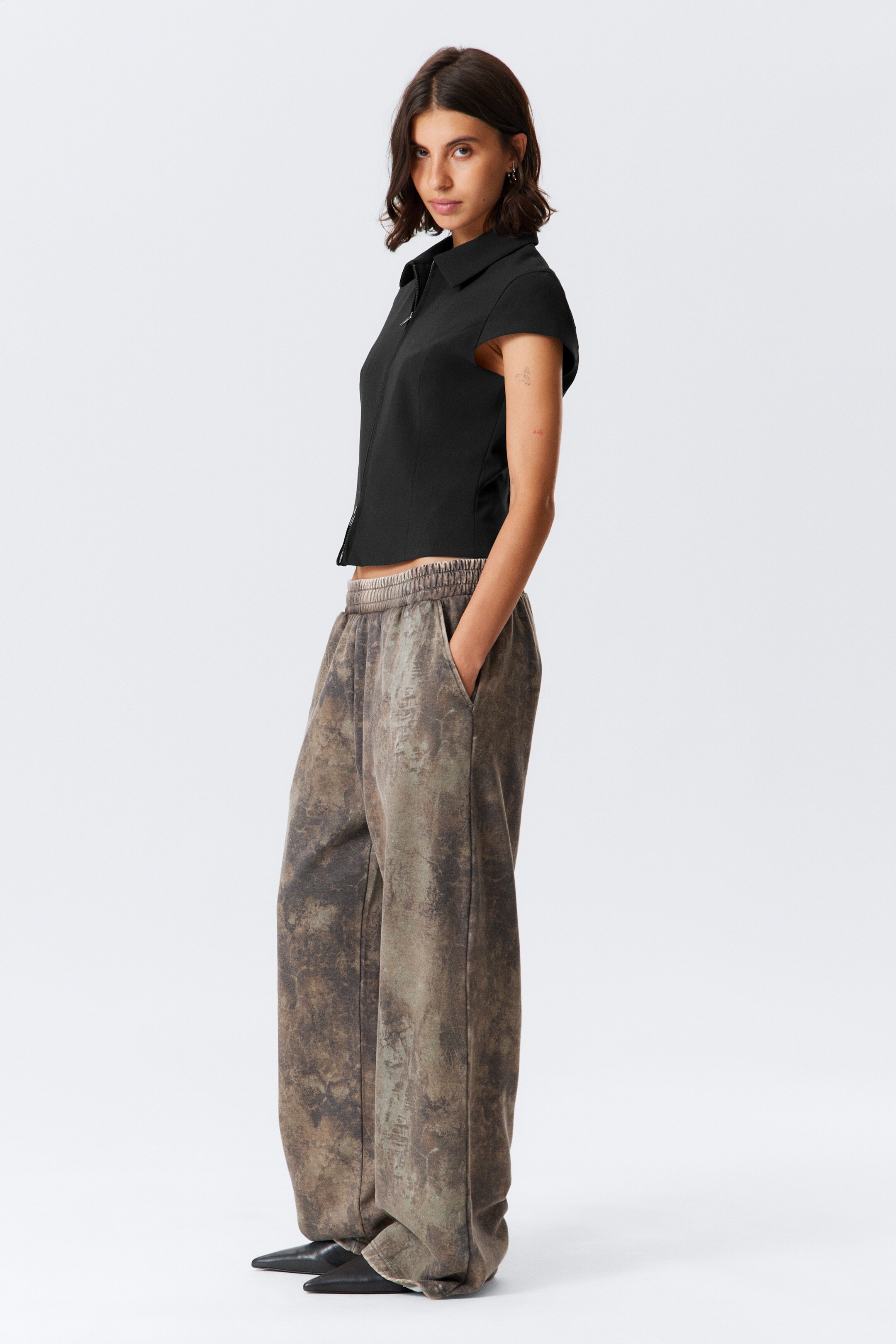 View larger image: A black short-sleeved top and patterned wide-leg pants are being presented. The top is featuring a collar and a front zipper closure, and is cropped at the waist. The pants are showcasing an elasticized waistband, side pockets, and a distressed, earthy-toned pattern, with their wide legs pooling at the ankles.