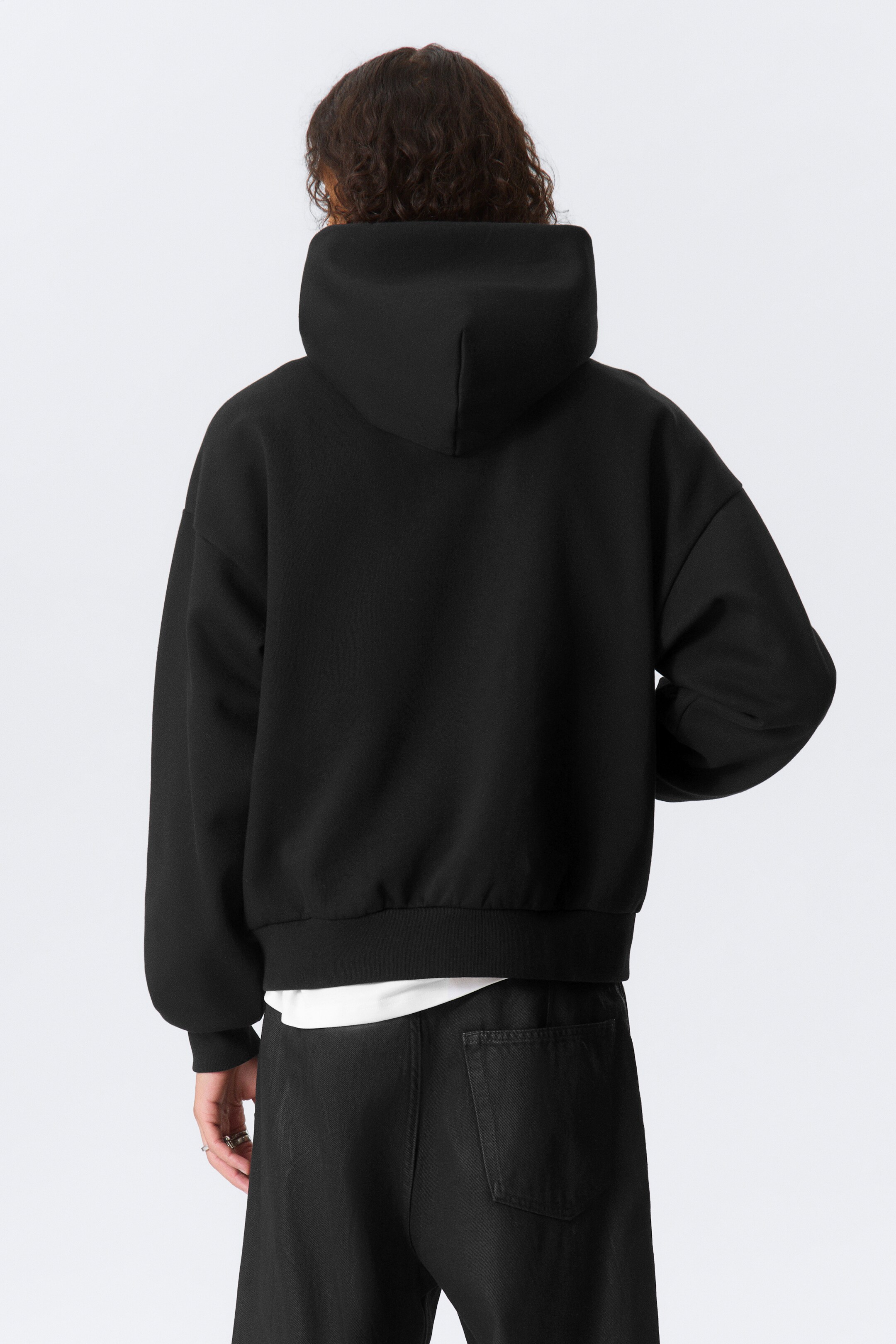 Grotere afbeelding bekijken: Een zwarte hoodie wordt gepresenteerd vanuit een achteraanzicht, met een grote capuchon, lange, loszittende mouwen met geribde manchetten en een geribde zoom. Een wit t-shirt wordt onder de zoom van de hoodie gedragen.