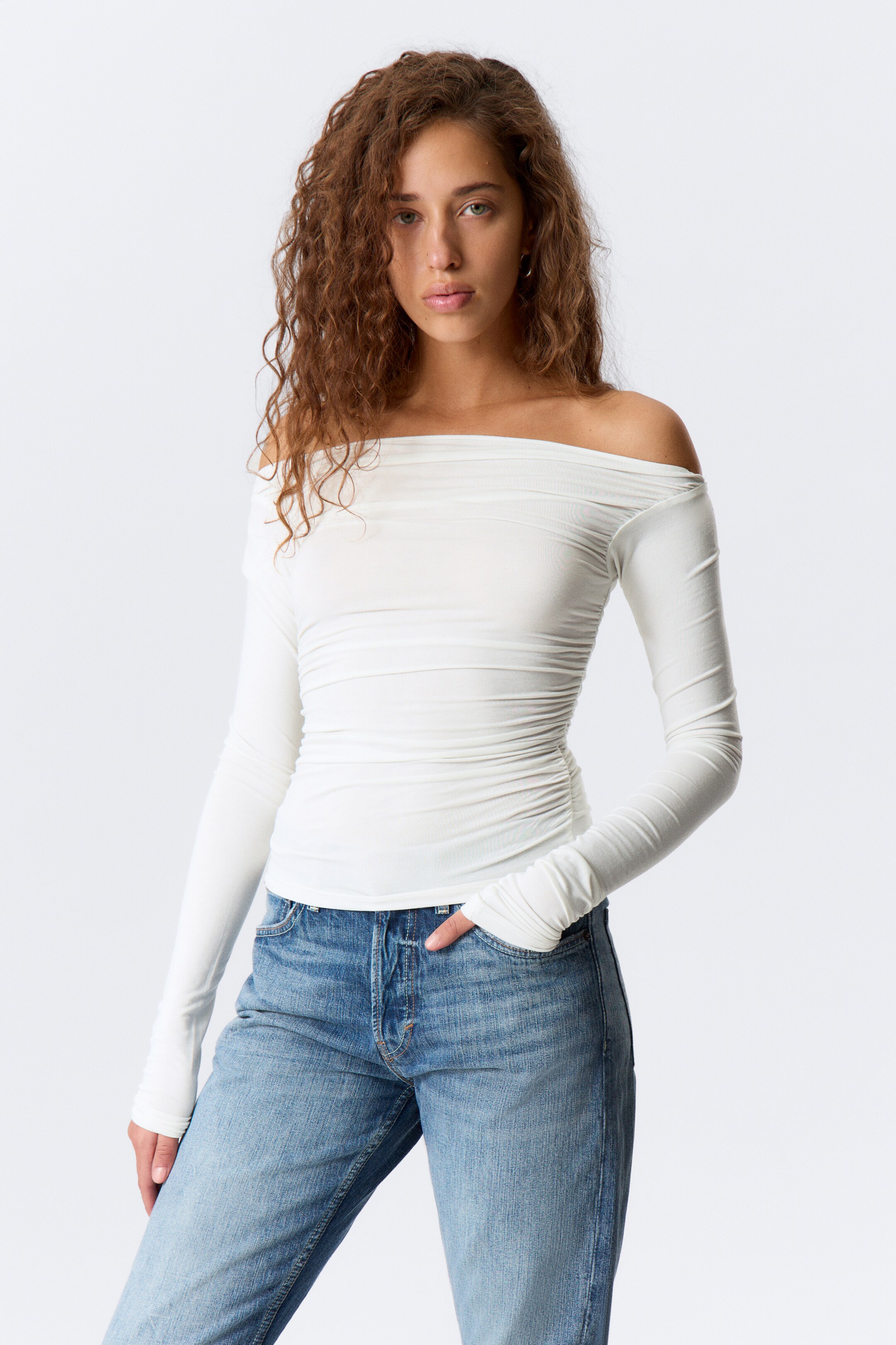 Agrandir l'image: Un haut blanc à épaules dénudées est présenté de face, avec un large décolleté drapé et des manches longues qui s'étendent sur les mains. Des fronces prononcées accentuent le torse, créant une silhouette ajustée.