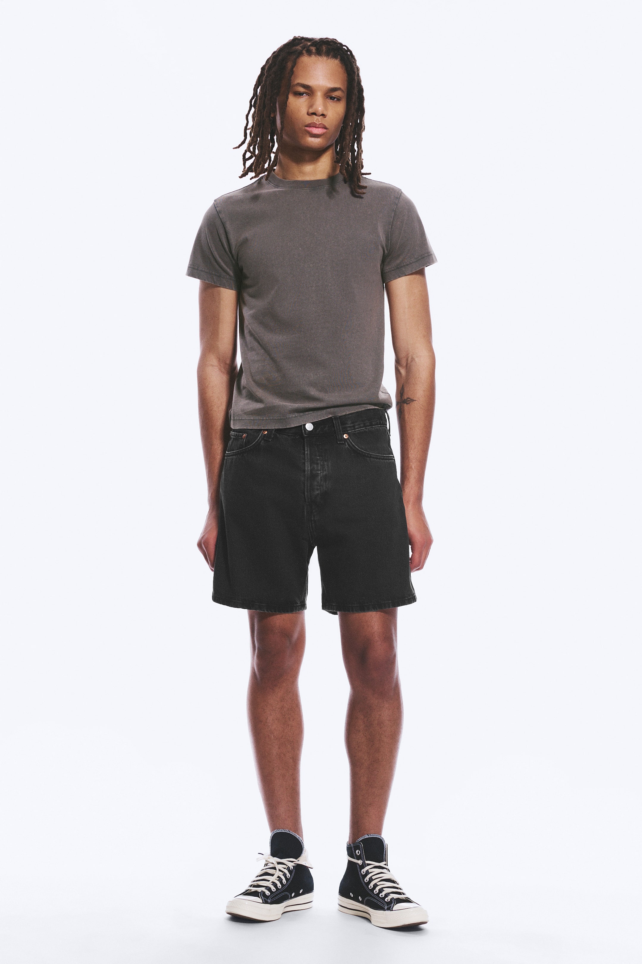Ver imagen más grande: Una camiseta de cuello redondo en gris oscuro con mangas cortas se lleva puesta, combinada con unos pantalones cortos de mezclilla negra con un dobladillo deshilachado y un ajuste relajado, presentada desde un ángulo frontal, y completada con zapatillas de lona negras de caña alta con cordones blancos.