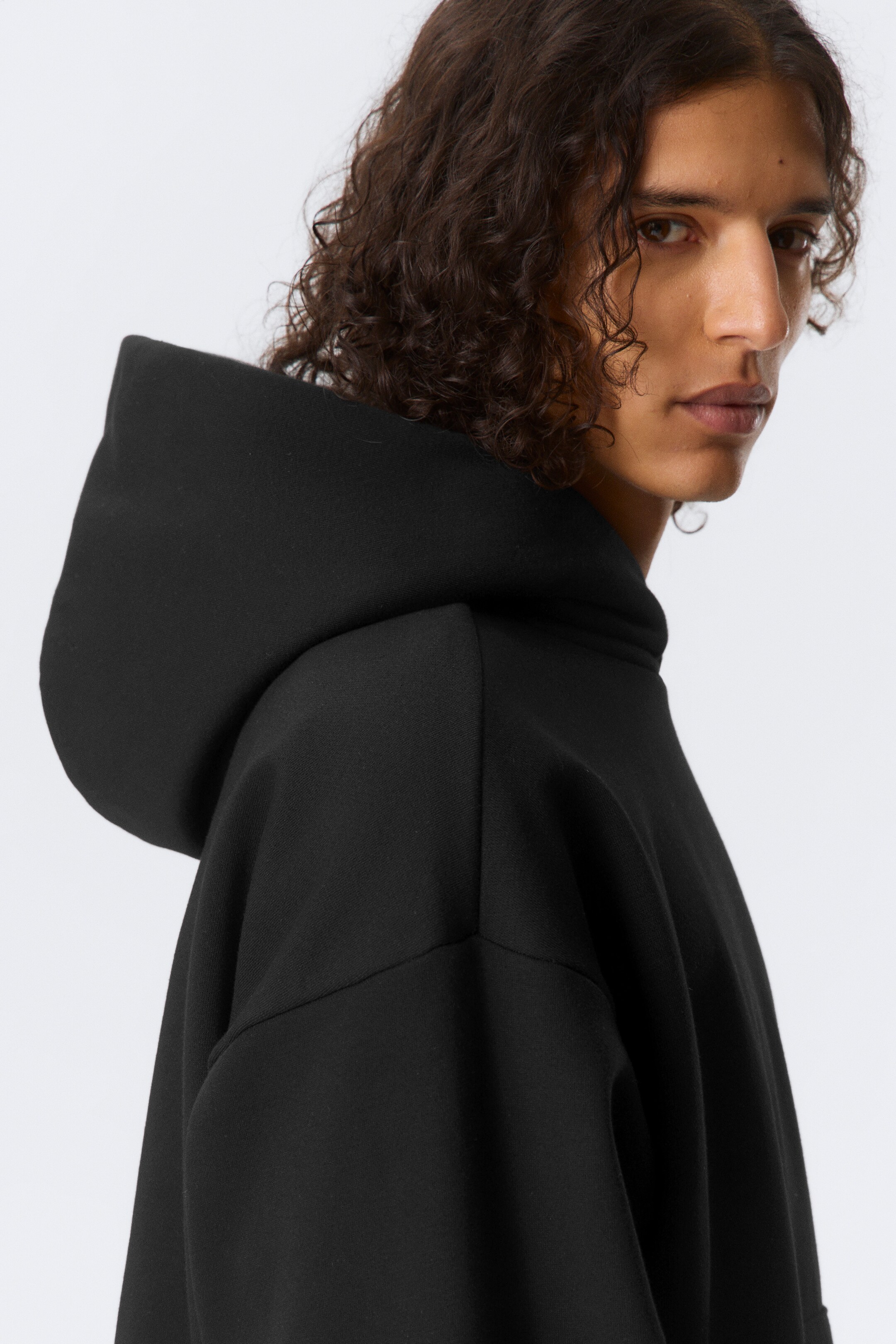 Grotere afbeelding bekijken: Een zwarte hoodie met een gevouwen capuchonconstructie en verlaagde schouders wordt gedragen door het model, waarbij de zachte katoenfleece losjes valt.