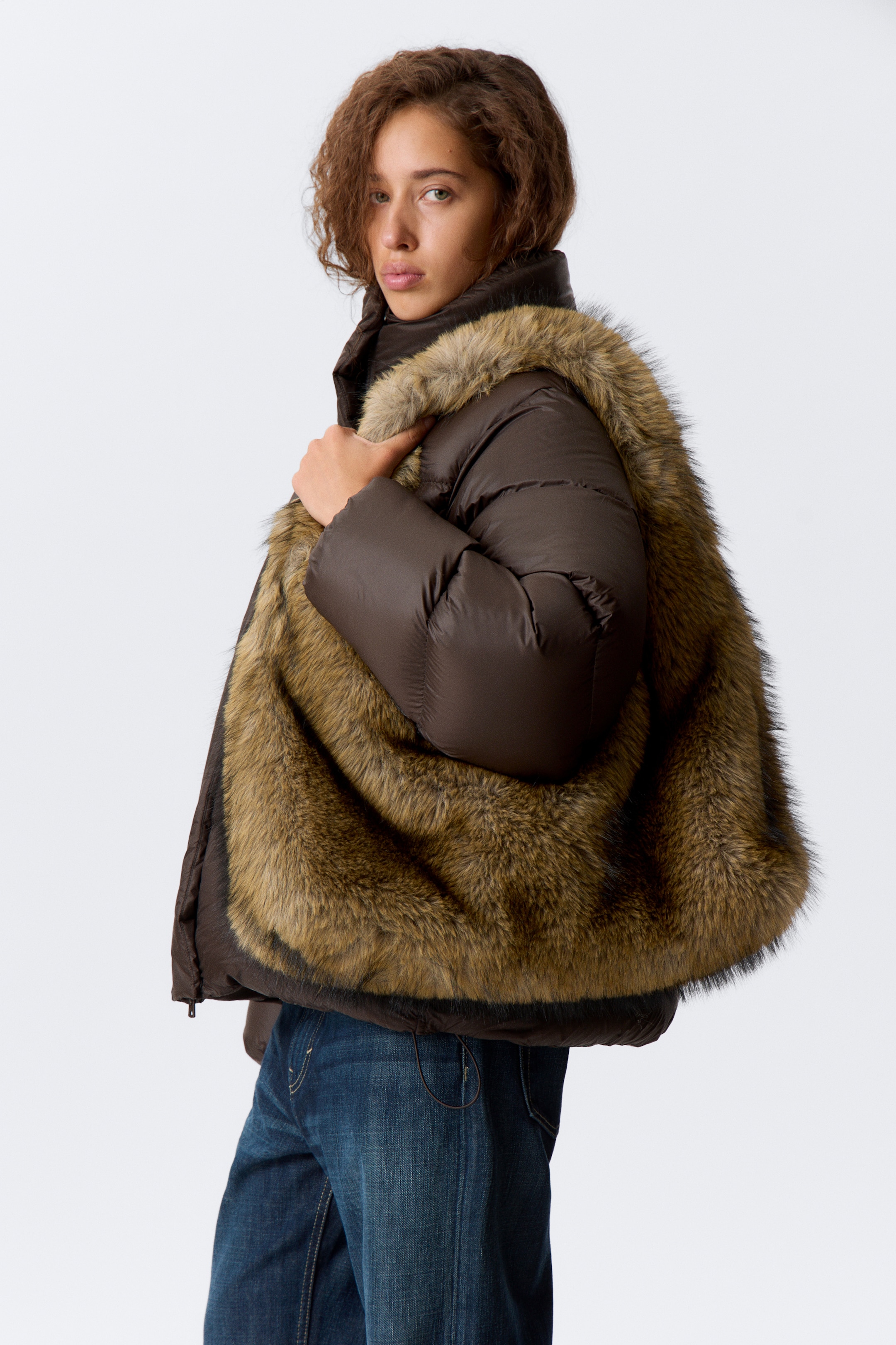 Größeres Bild ansehen: Eine braune wattierte Jacke wird präsentiert, leicht nach rechts geneigt, wobei das zottelige, mehrfarbige braune Kunstfellfutter deutlich zu sehen ist. Die Jacke hat einen hohen Kragen und wattierte lange Ärmel.