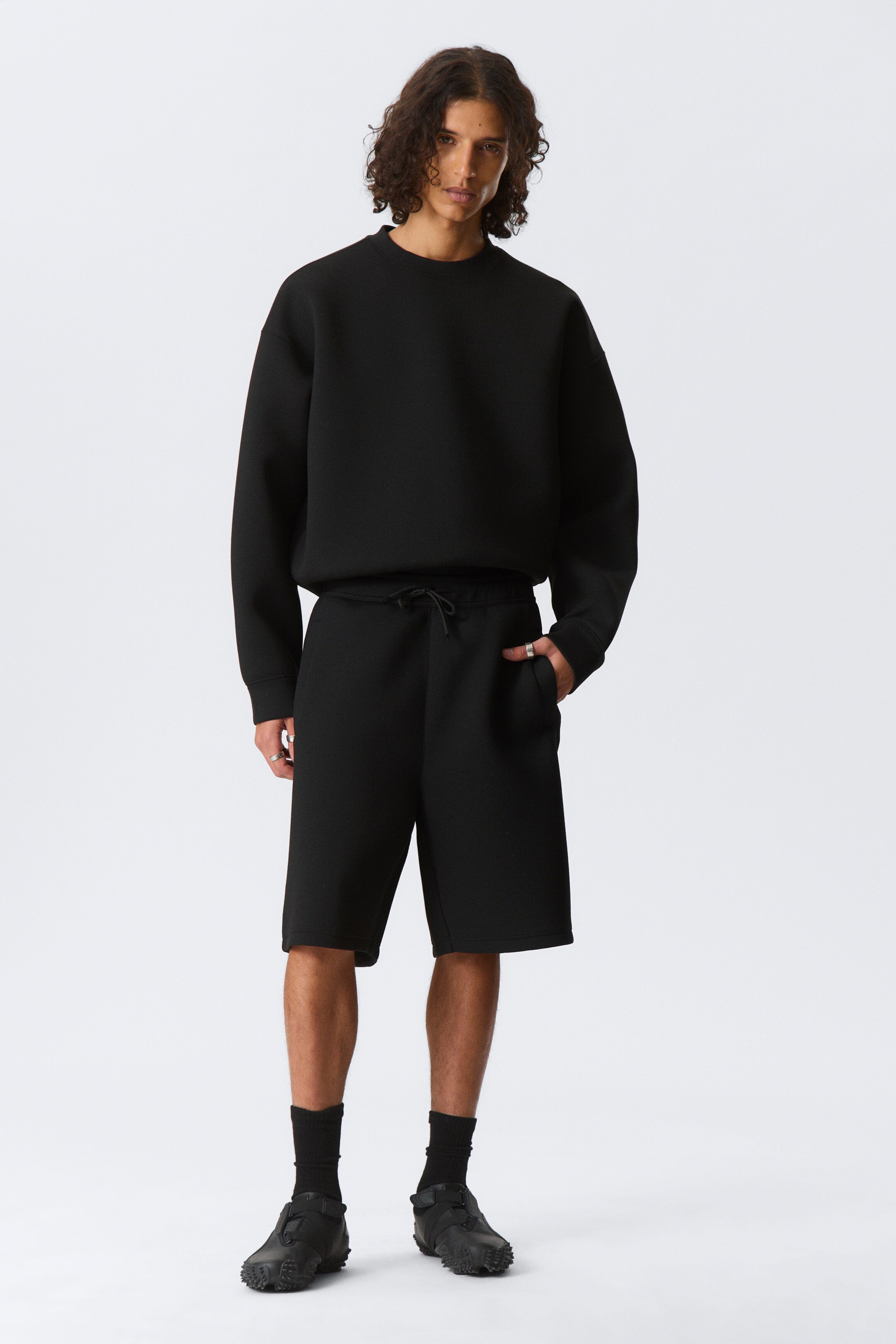 Vis større bilde: En svart crewneck-genser med tekstur og avslappet passform, sammen med matchende svarte shorts med tekstur og snøring, presenteres fra en frontvendt vinkel.
