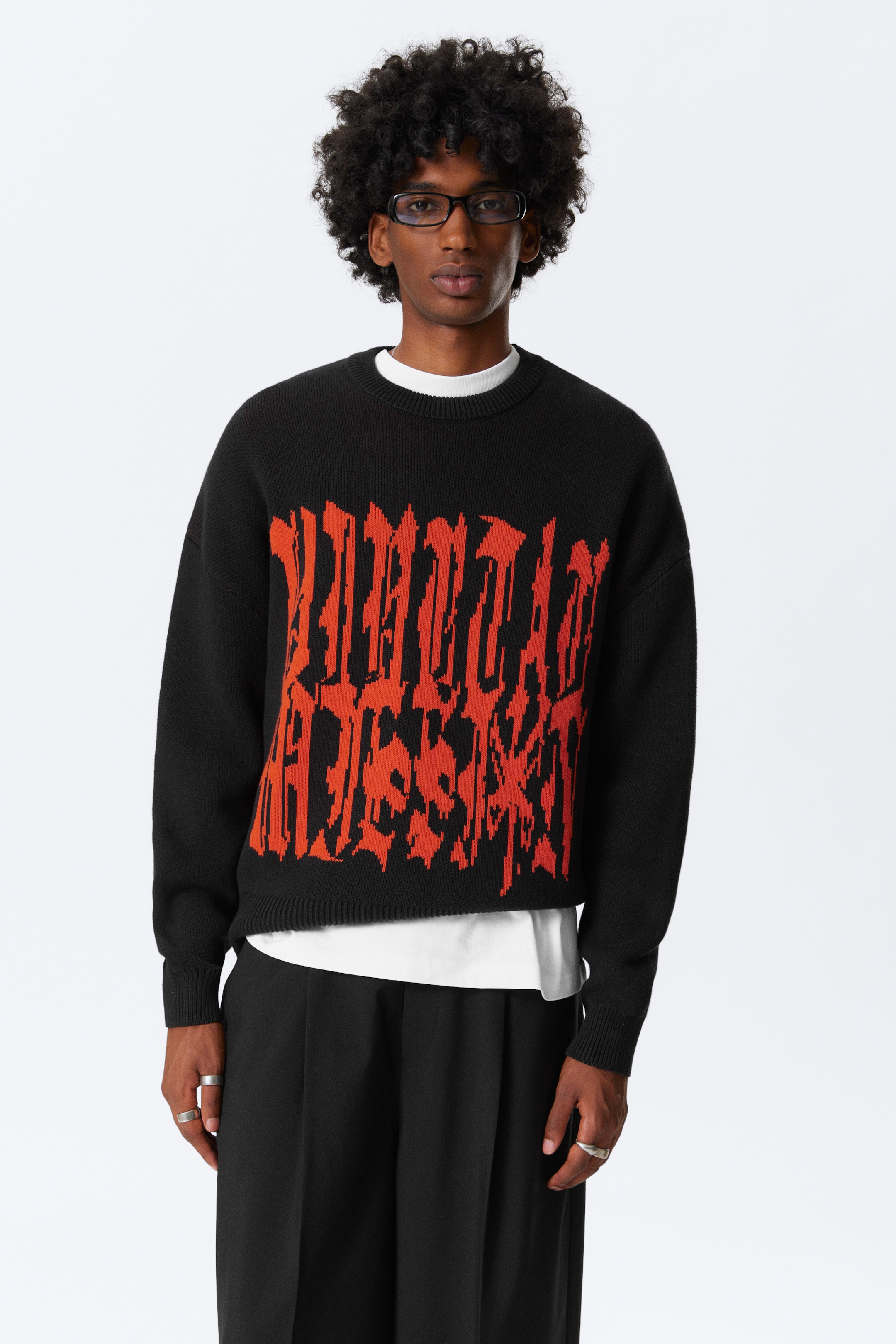 Agrandir l'image: Un pull à col rond noir est porté, avec un grand design de texte graphique orange sur la poitrine. Le pull présente des poignets et un ourlet côtelés, et est superposé à un t-shirt blanc visible au niveau du cou et de l'ourlet.