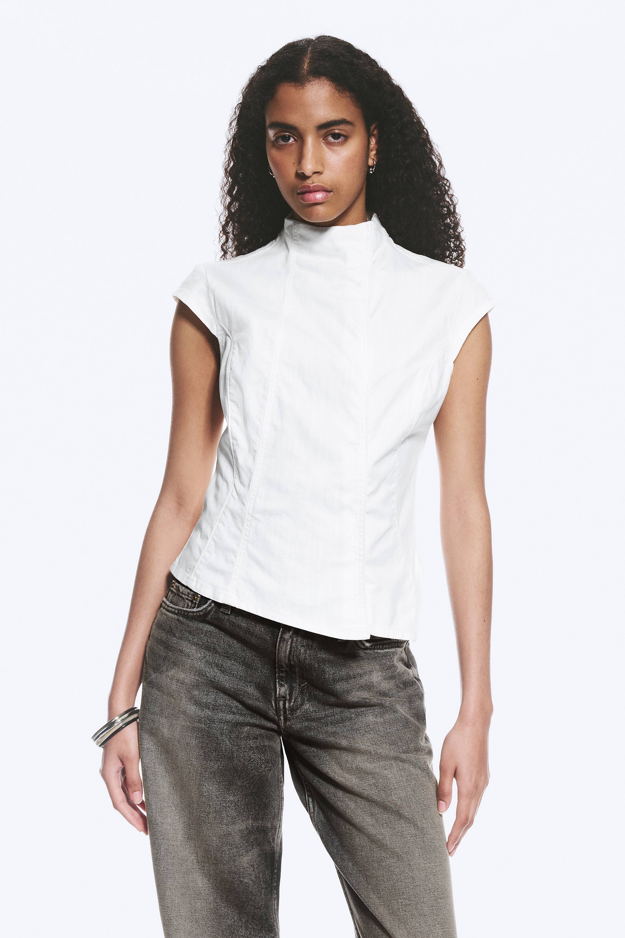 Ingrandisci l'immagine: Un top bianco senza maniche presenta un colletto rialzato e maniche a cappuccio. Mostra cuciture verticali su misura sul davanti, creando una silhouette aderente.