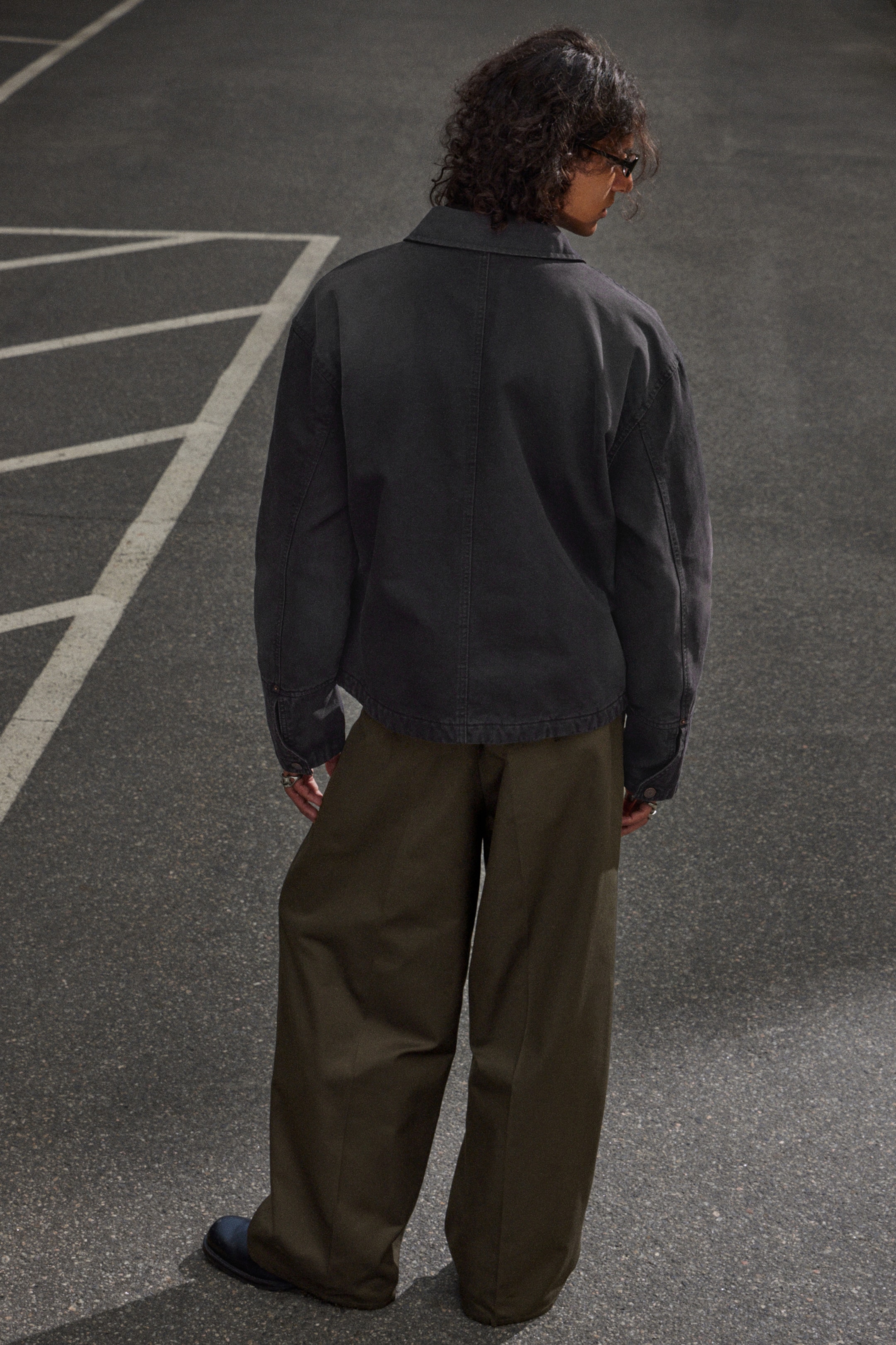 View larger image: A charcoal gray jacket is being worn, shown from the back, featuring a classic collar, prominent vertical seams, and a relaxed fit. It is being paired with wide-leg olive green trousers that are also visible from the back, draping loosely and pooling at the ankles.
