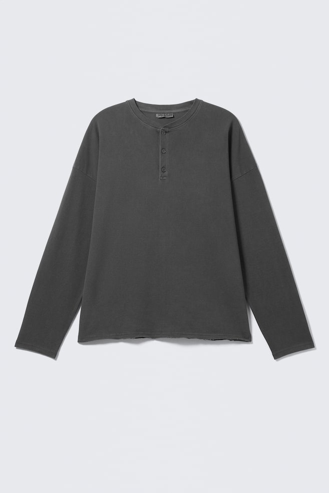 Ein dunkelgraues Langarm-Henley-Shirt liegt flach und zeigt nach vorne. Es verfügt über eine Drei-Knopf-Leiste, überschnittene Schultern und einen ungesäumten, rohen Saum. - 1