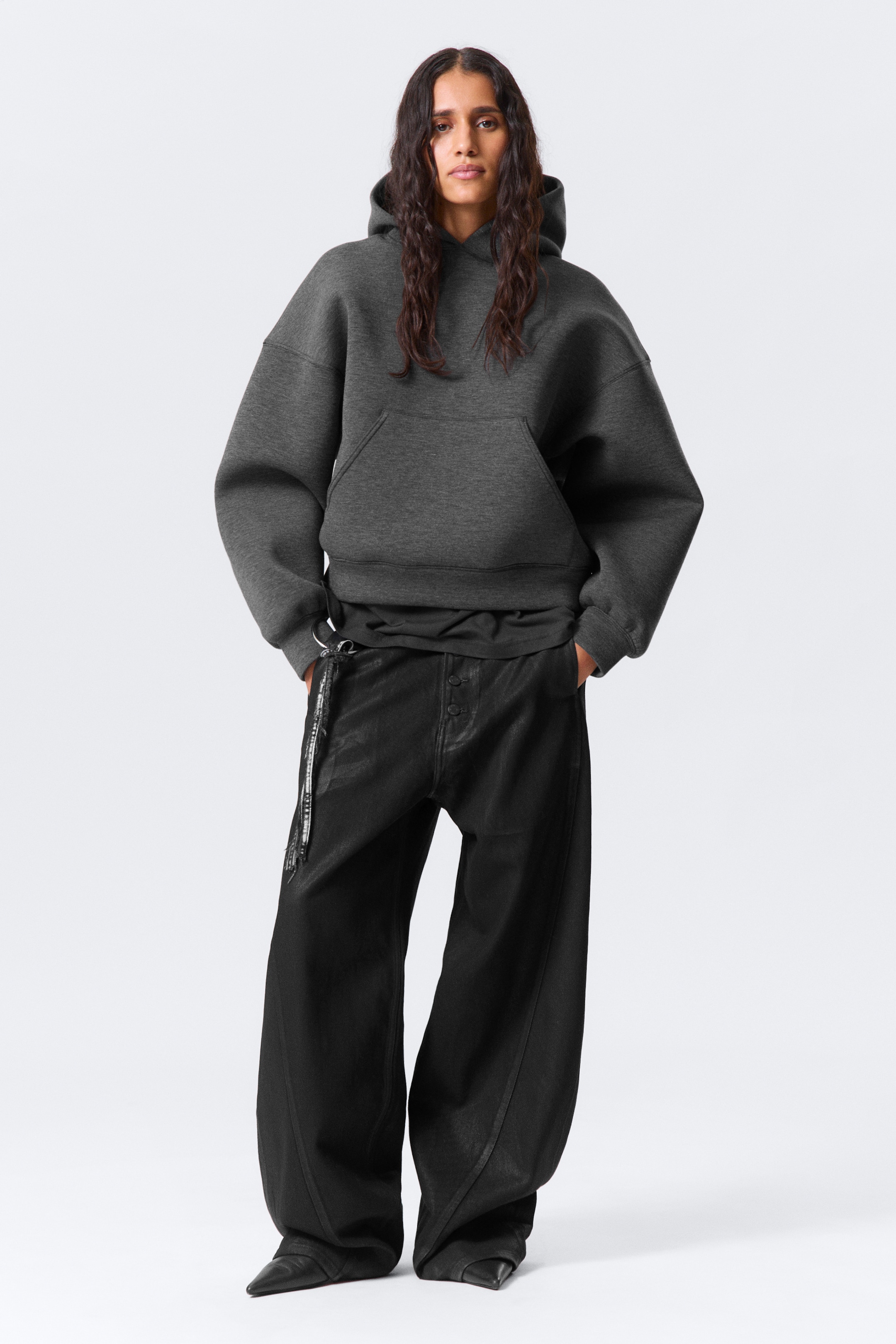 Agrandir l'image: Une veste à capuche en néoprène gris foncé, taillée pour une coupe ample, est portée par le mannequin, avec une capuche spacieuse, des épaules tombantes, une poche kangourou, et des poignets et une ligne d'ourlet lisses. Les mains du mannequin reposent dans la poche kangourou.