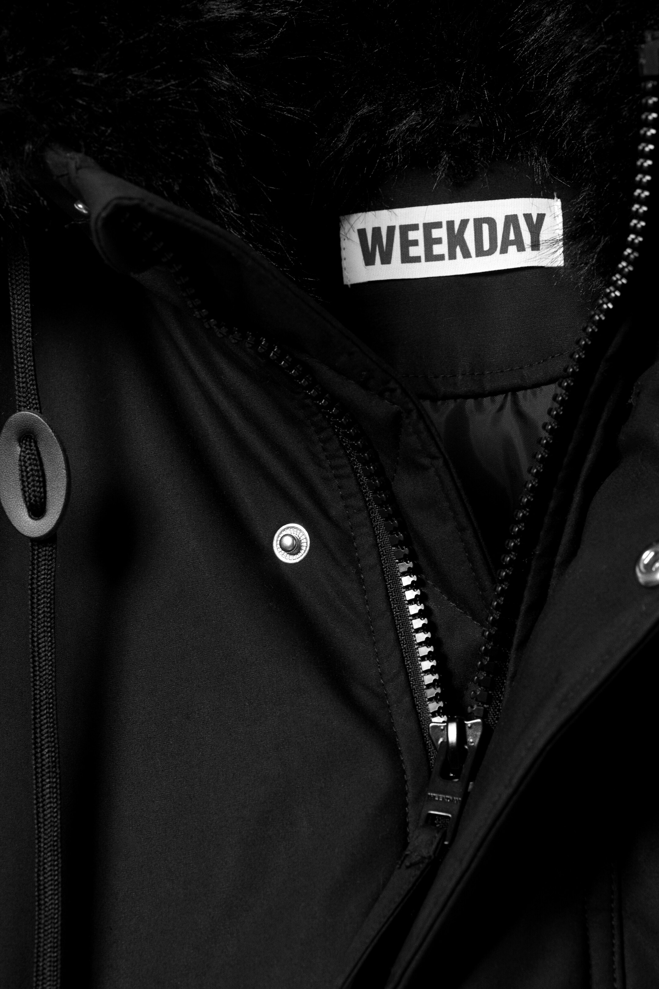 Grotere afbeelding bekijken: Een zwarte parka met een zwarte faux fur capuchonrand, een witte "WEEKDAY" merklabel aan de binnenkant van de halslijn, en een gedeeltelijk geopende voorrits met een zilveren trekker. Zilveren drukknoopjes sieren de sluiting, en een zwart trekkoord met een stopper is zichtbaar aan de linkerkant.