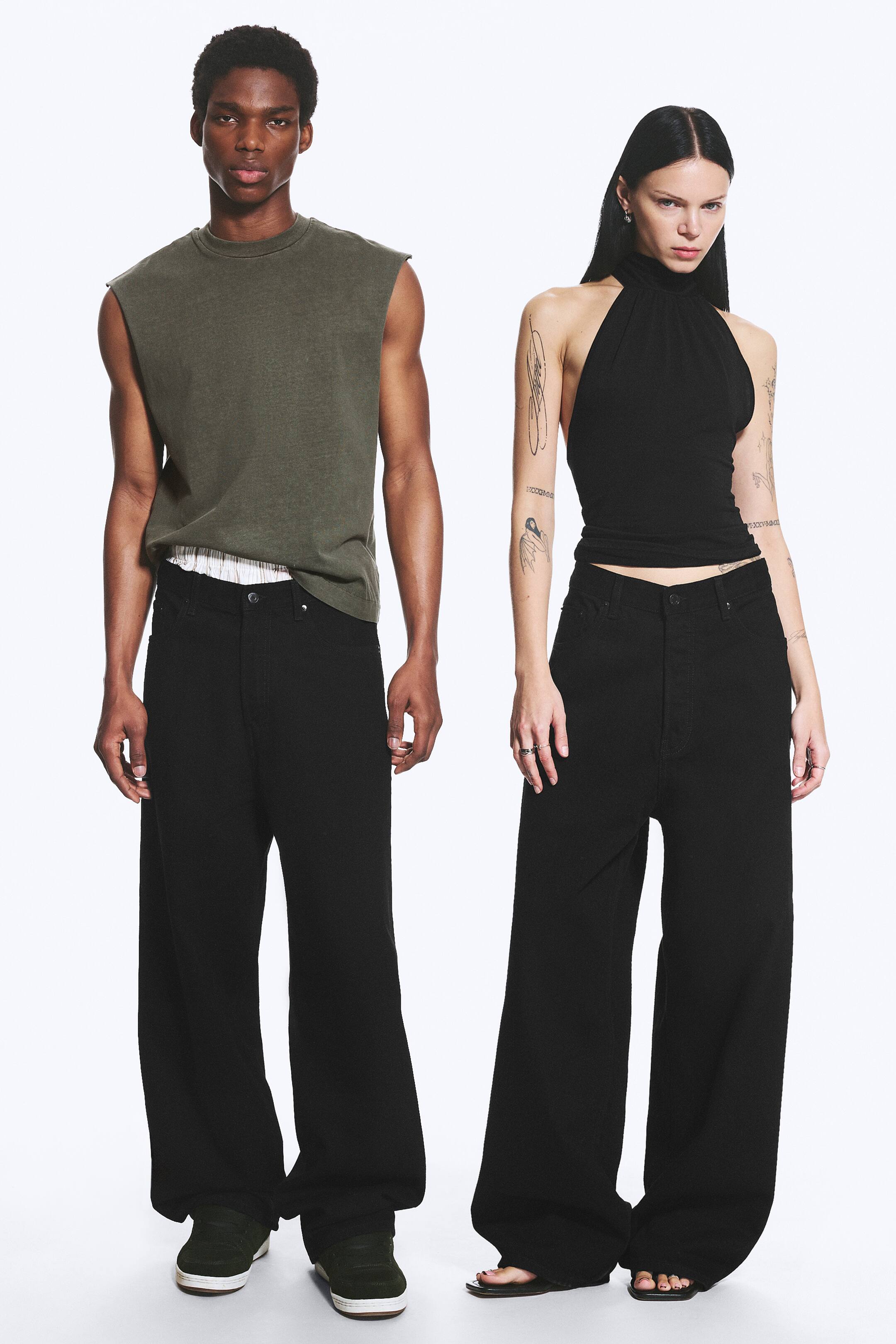 Zobacz większy obraz: Czarne spodnie z wysokim stanem prezentowane są na dwóch modelkach, stojących przodem. Spodnie charakteryzują się luźnym, szerokim krojem nogawek, sięgającym do podłogi, z widocznym zapięciem na guzik i zamek błyskawiczny w talii.