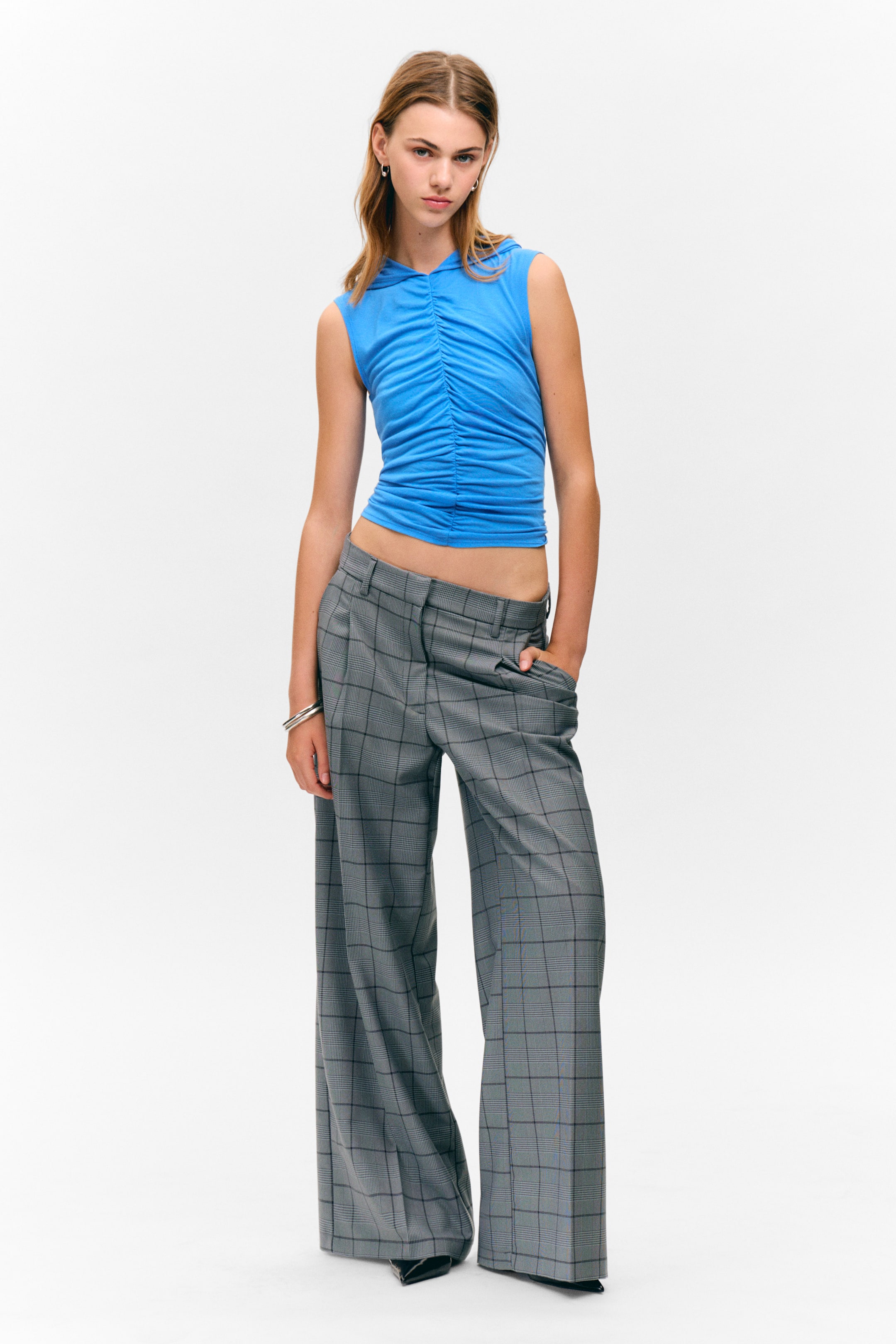 Zobraziť väčší obrázok: Modrý bezrukávový top s kapucňou je charakteristický rozsiahlym vertikálnym riasením na prednej strane, pôsobiac skráteným do pásu. Je kombinovaný so sivými širokými nohavicami, ktoré majú výrazný károvaný vzor, záhyby v páse a bočné vrecká.