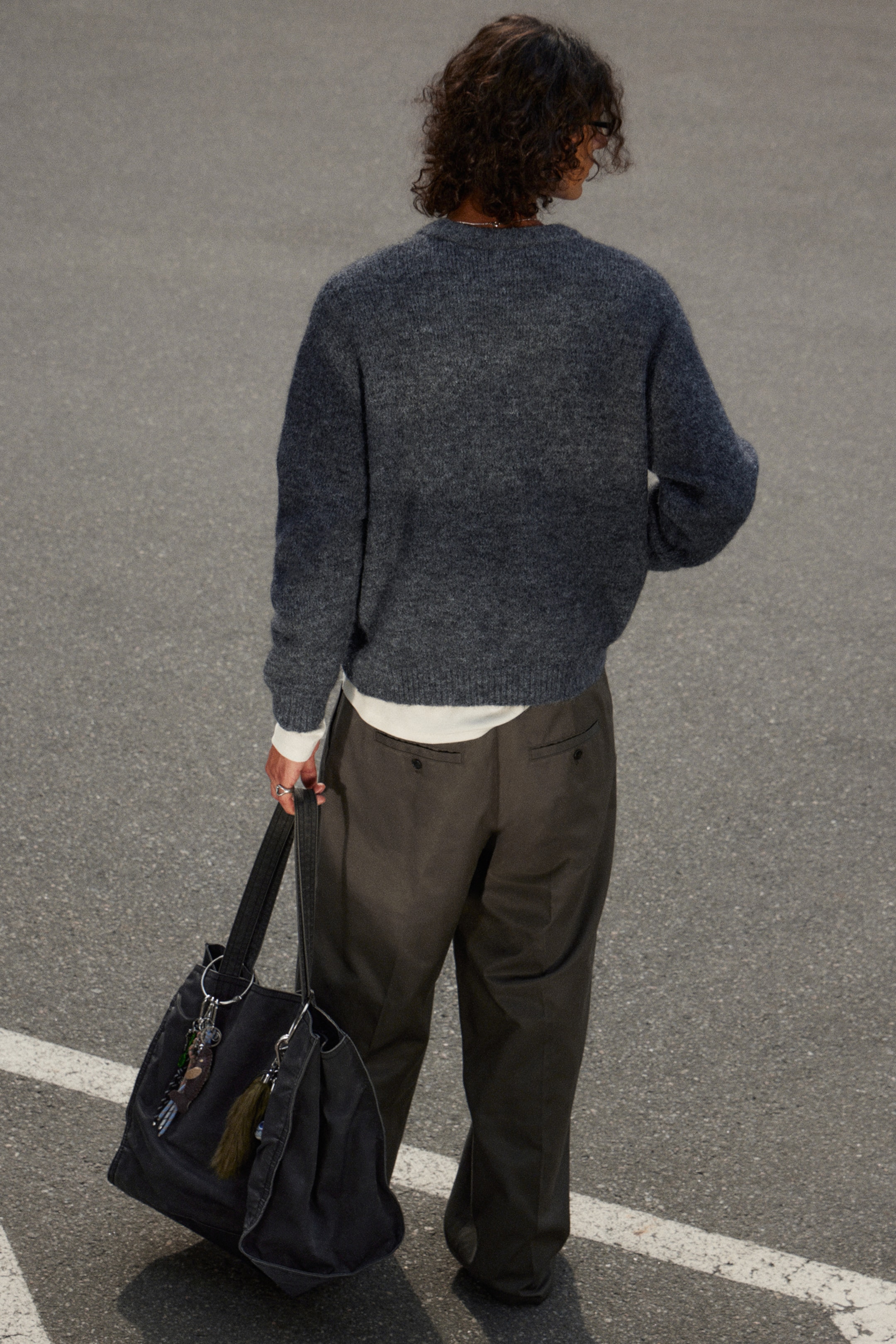Grösseres Bild ansehen: Ein dunkelgrauer, flauschiger Strickpullover mit Rundhalsausschnitt wird aus einem hinteren Winkel getragen, über einem weißen langärmeligen Hemd, das an den Manschetten und am Saum sichtbar ist. Dunkelbraune, locker sitzende Hosen mit geknöpften Gesäßtaschen werden ebenfalls getragen, während eine große dunkelgraue Canvas-Tragetasche, verziert mit einem runden Schlüsselring, einem Perlenanhänger und einer Quaste, an der Seite der Person gehalten wird.