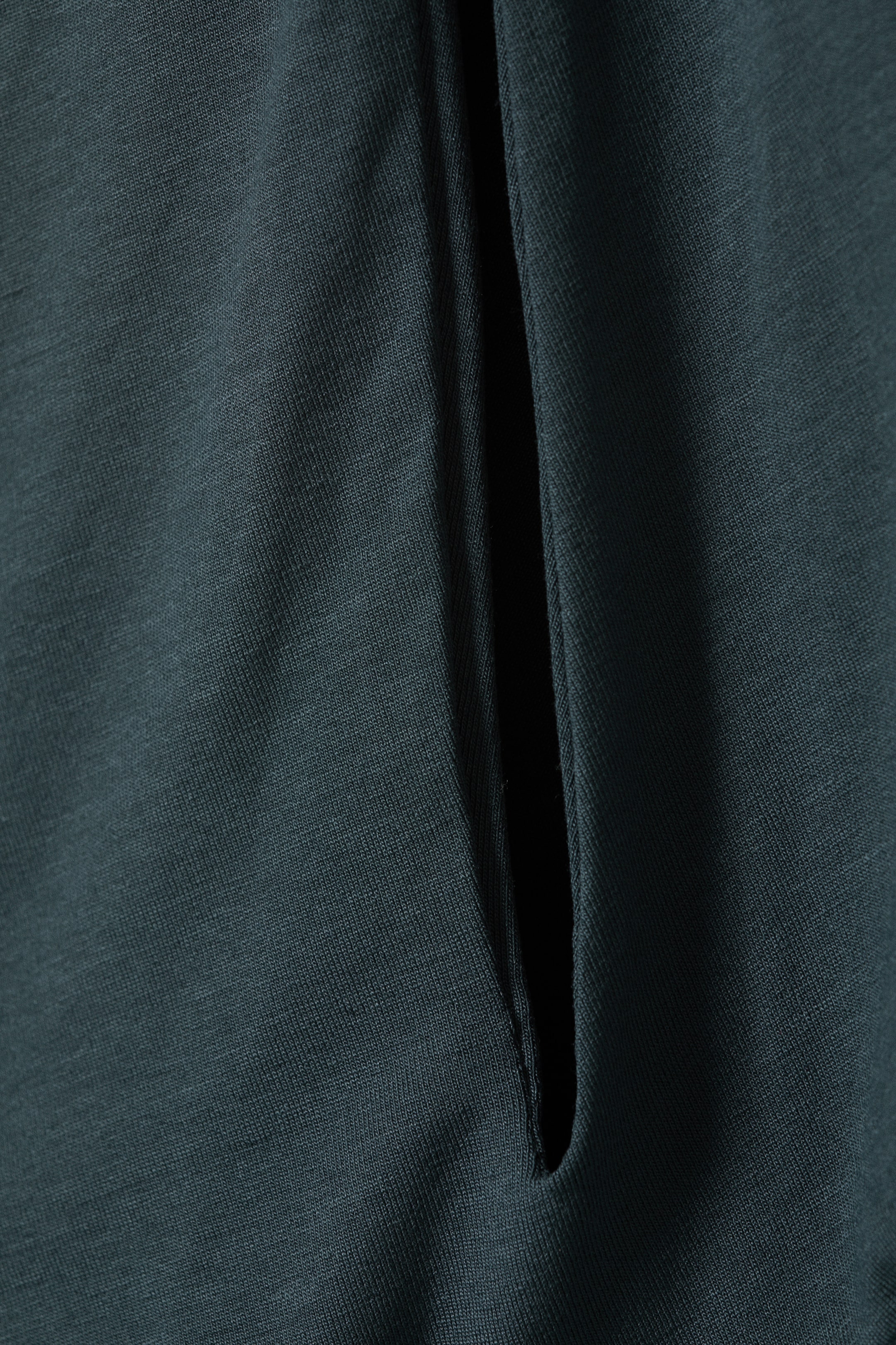 Grösseres Bild ansehen: Ein regulär geschnittenes, einseitiges Oberteil in dunklem Teal verfügt über ein vertikales Front-Cut-Out-Detail. Die glatten Kanten des Cut-Outs enthüllen eine doppellagige Vorderseite, ohne dass eine sichtbare Steppnaht auf dem Stoff zu sehen ist.