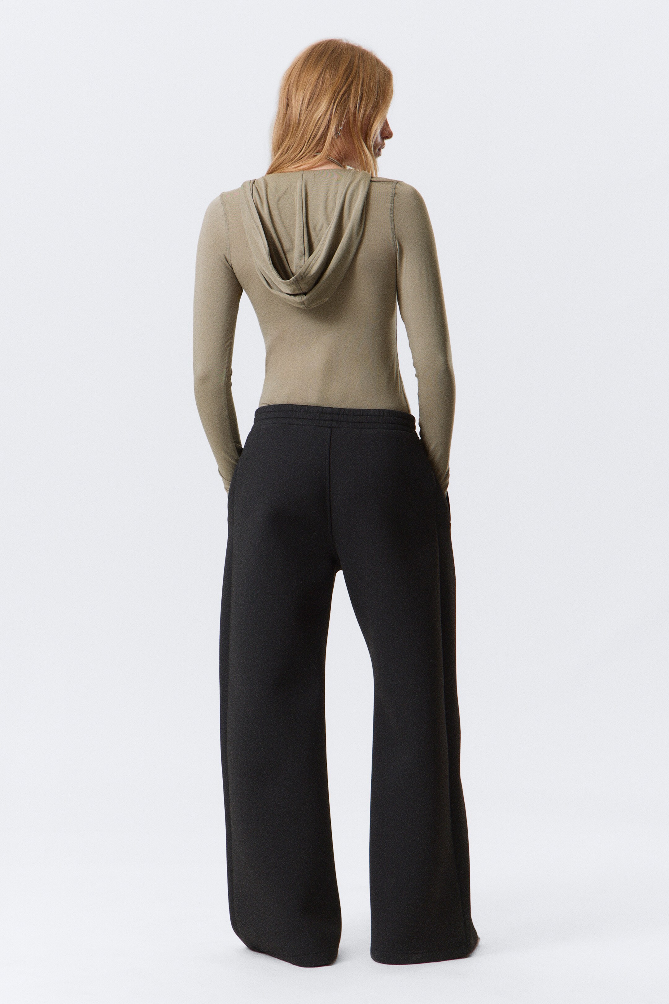 Ver imagen más grande: Se muestra a una modelo de espaldas, vistiendo un top ajustado de manga larga con capucha en color verde oliva, con la capucha cayendo suavemente, y pantalones negros de pierna ancha que fluyen holgadamente desde las caderas.