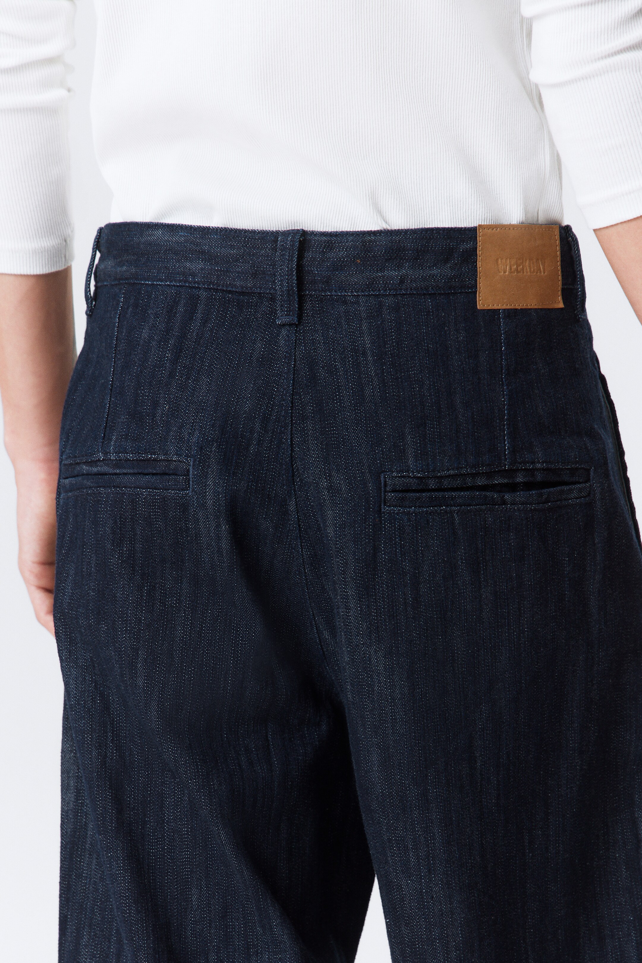 Agrandir l'image: Un pantalon en jean indigo foncé est vu de dos, avec une ceinture ornée de passants et une étiquette marron en relief avec l'inscription 'WEEKDAY' à l'arrière droit. Le pantalon présente deux poches arrière passepoilées horizontales et un tissage en denim distinctif, avec une chemise blanche côtelée rentrée dans la ceinture.