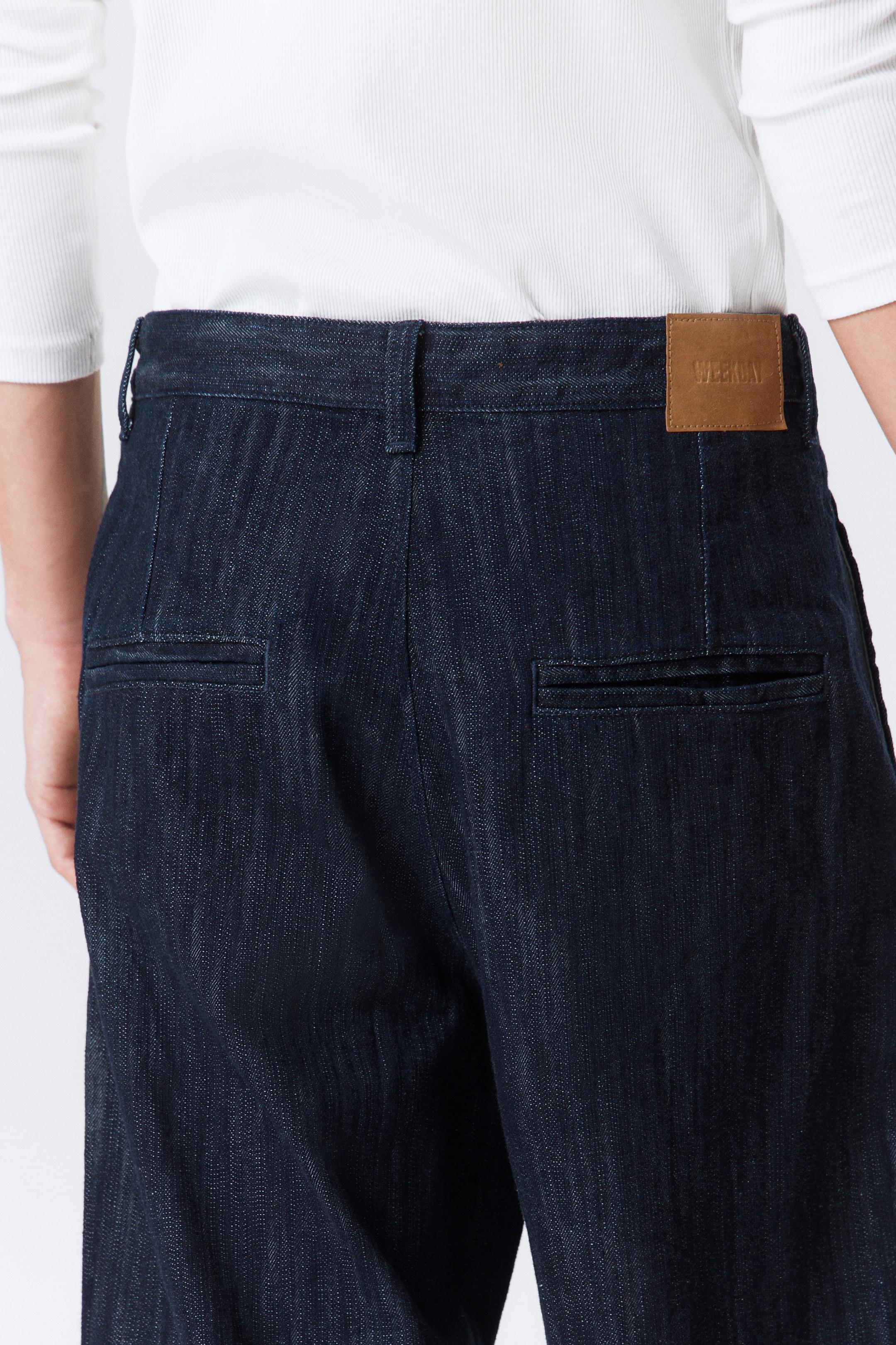 Ver imagen más grande: Pantalones de mezclilla índigo oscuro se ven desde atrás, con una pretina con trabillas para cinturón y una etiqueta de parche marrón con relieve 'WEEKDAY' en la parte trasera derecha. Los pantalones presentan dos bolsillos traseros horizontales de ojal y un tejido de mezclilla distintivo, con una camisa blanca de canalé metida en la pretina.