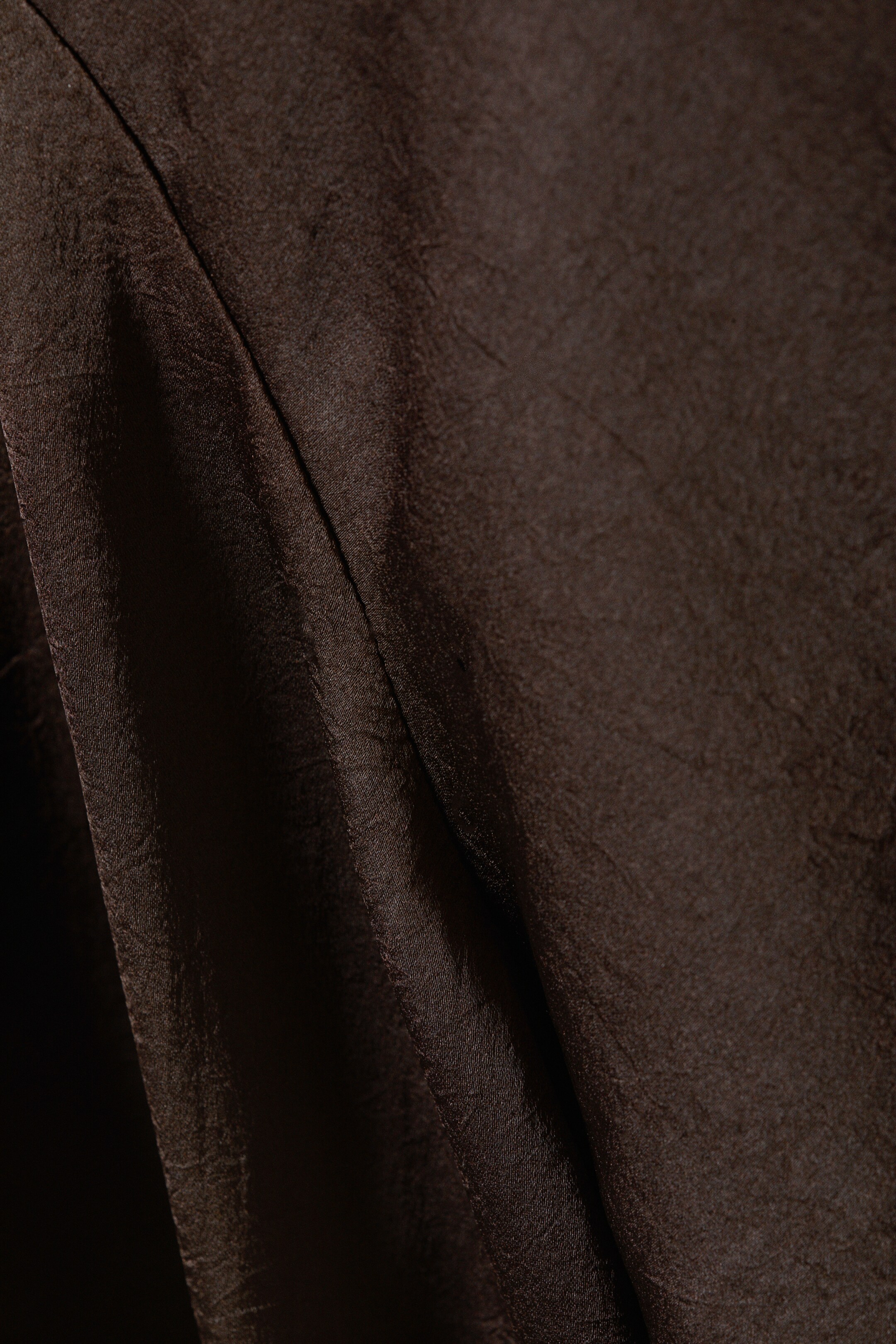 View larger image: A dark brown fabric is filling the frame, exhibiting a subtle sheen and a distinct crinkled texture. A prominent vertical seam is running down the left, creating soft drapes and folds.