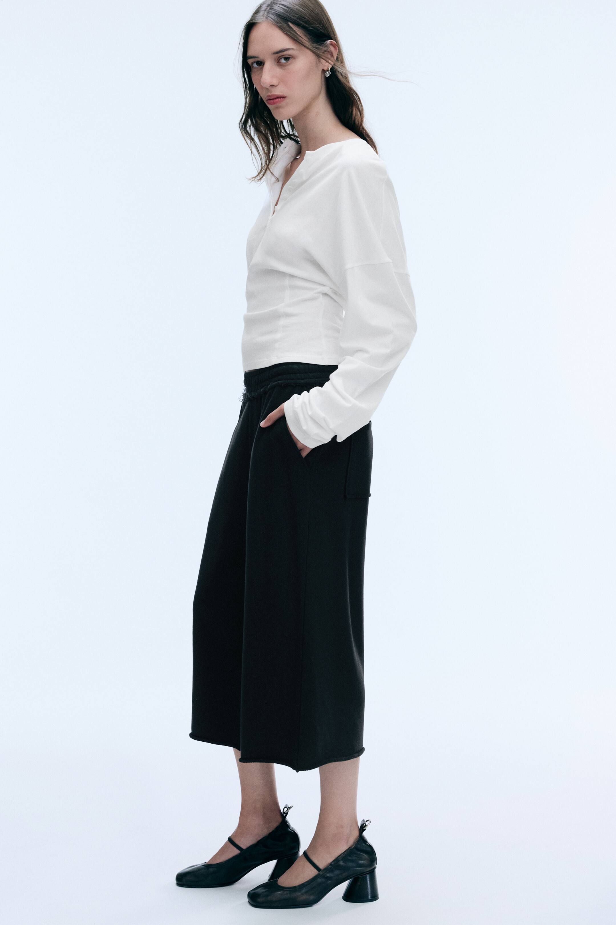 Visualizza immagine più grande: Pantaloni neri in spugna 7/8 con silhouette a gamba ampia, vita elasticizzata con dettagli a taglio vivo e tasche laterali, indossati dalla modella. La modella è in piedi di profilo con una mano appoggiata nella tasca del pantalone, abbinando i pantaloni a un top bianco a maniche lunghe e scarpe nere con tacco.