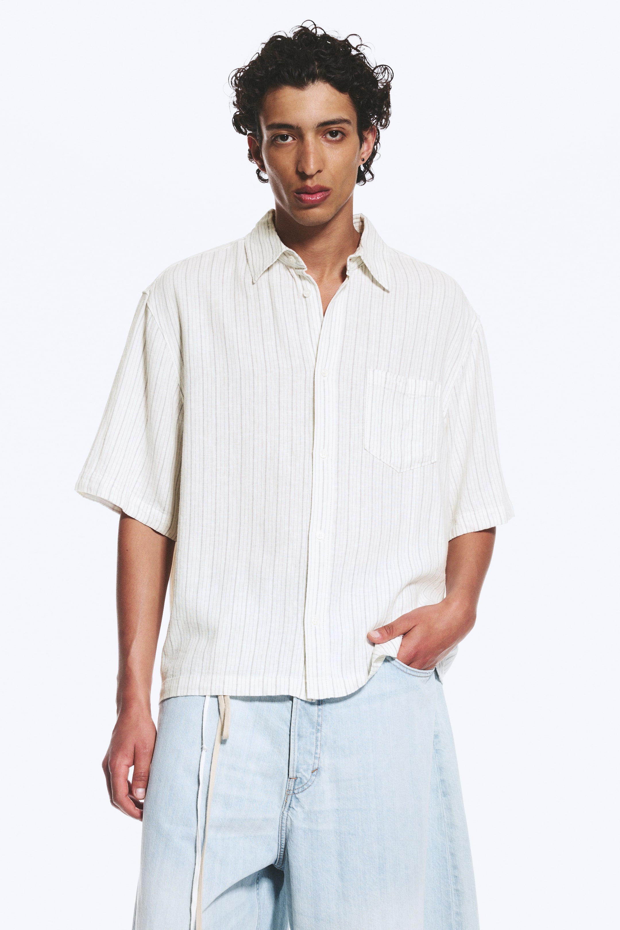 Se større billede: En off-white, kortærmet skjorte med diskrete lodrette nålestriber. Den har en spids krave, en ubesluttet knaplukning foran med lyse knapper og en enkelt brystlomme på venstre side, hvilket giver en løs pasform.