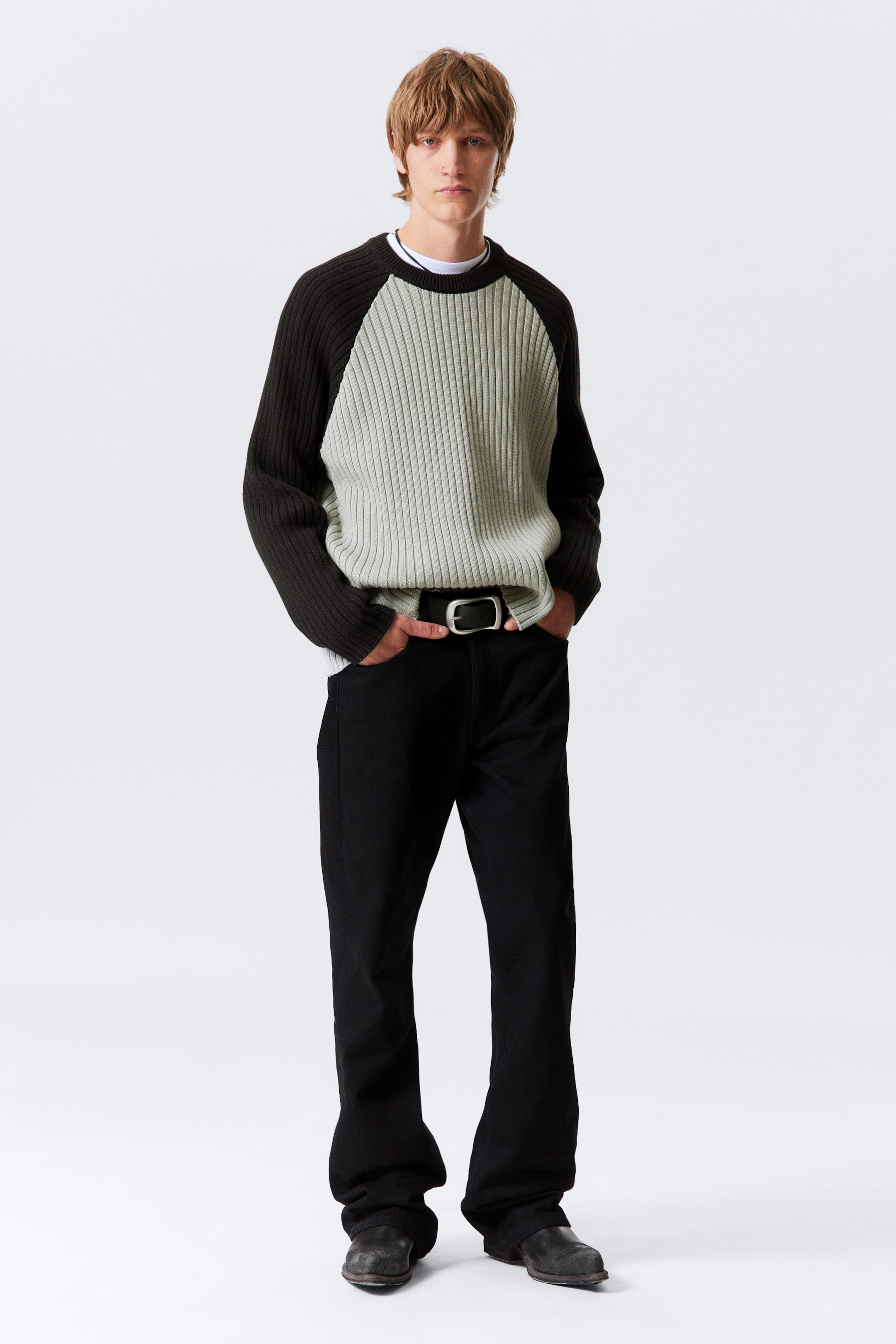 Größeres Bild anzeigen: Ein kastiger, zweifarbiger, breiter Rippstrickpullover mit einem hellgrünen Körper und dunklen Raglanärmeln wird vom Model über einem weißen Unterhemd getragen. Das Model steht mit den Händen in den Taschen.